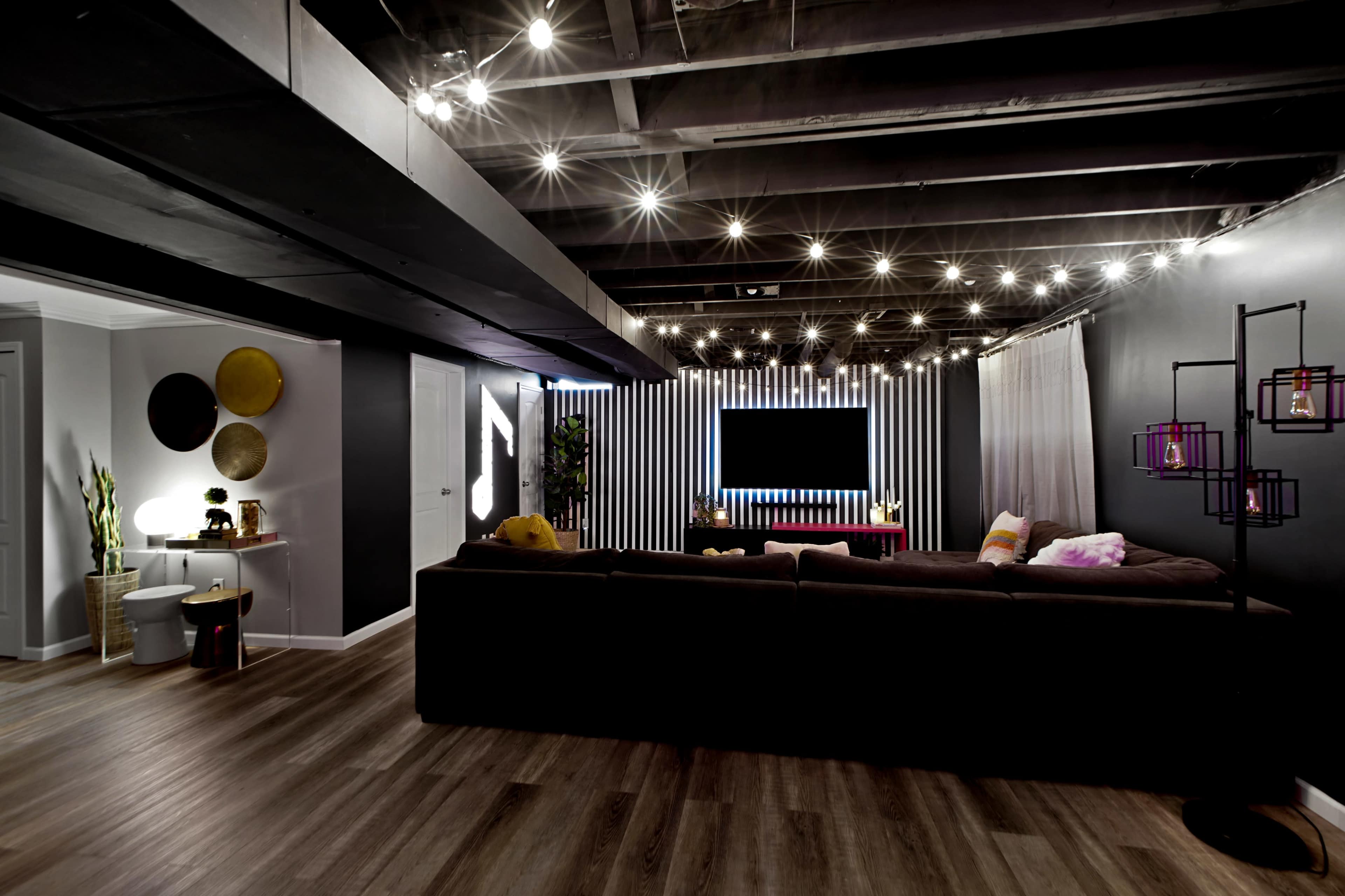 The image shows a modern living space with a sectional couch, decorative lighting, and a wall-mounted TV surrounded by vertical stripes.