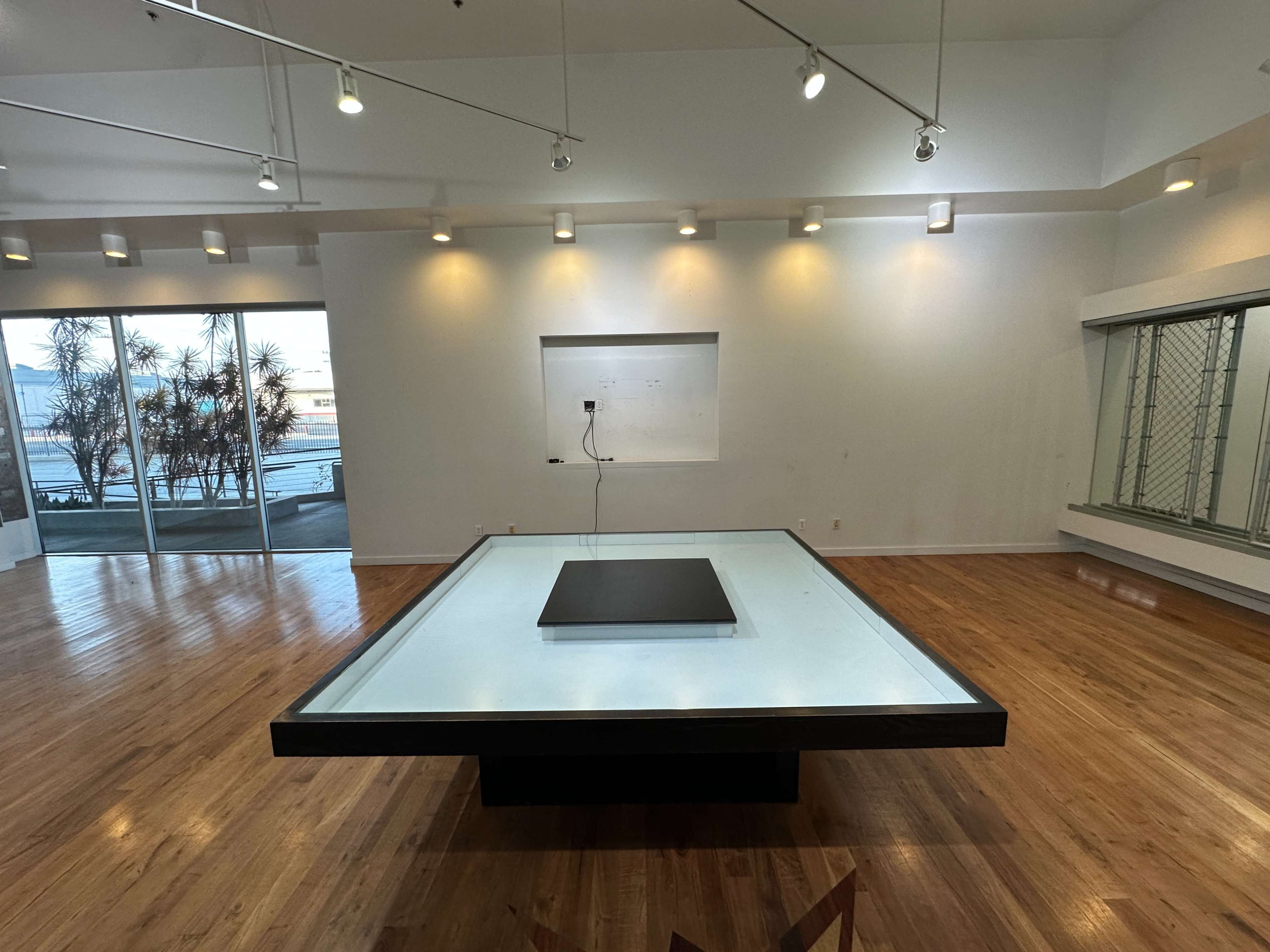 The image shows a minimalist room with wooden flooring, featuring a large, illuminated table in the center and windows displaying outdoor plants.