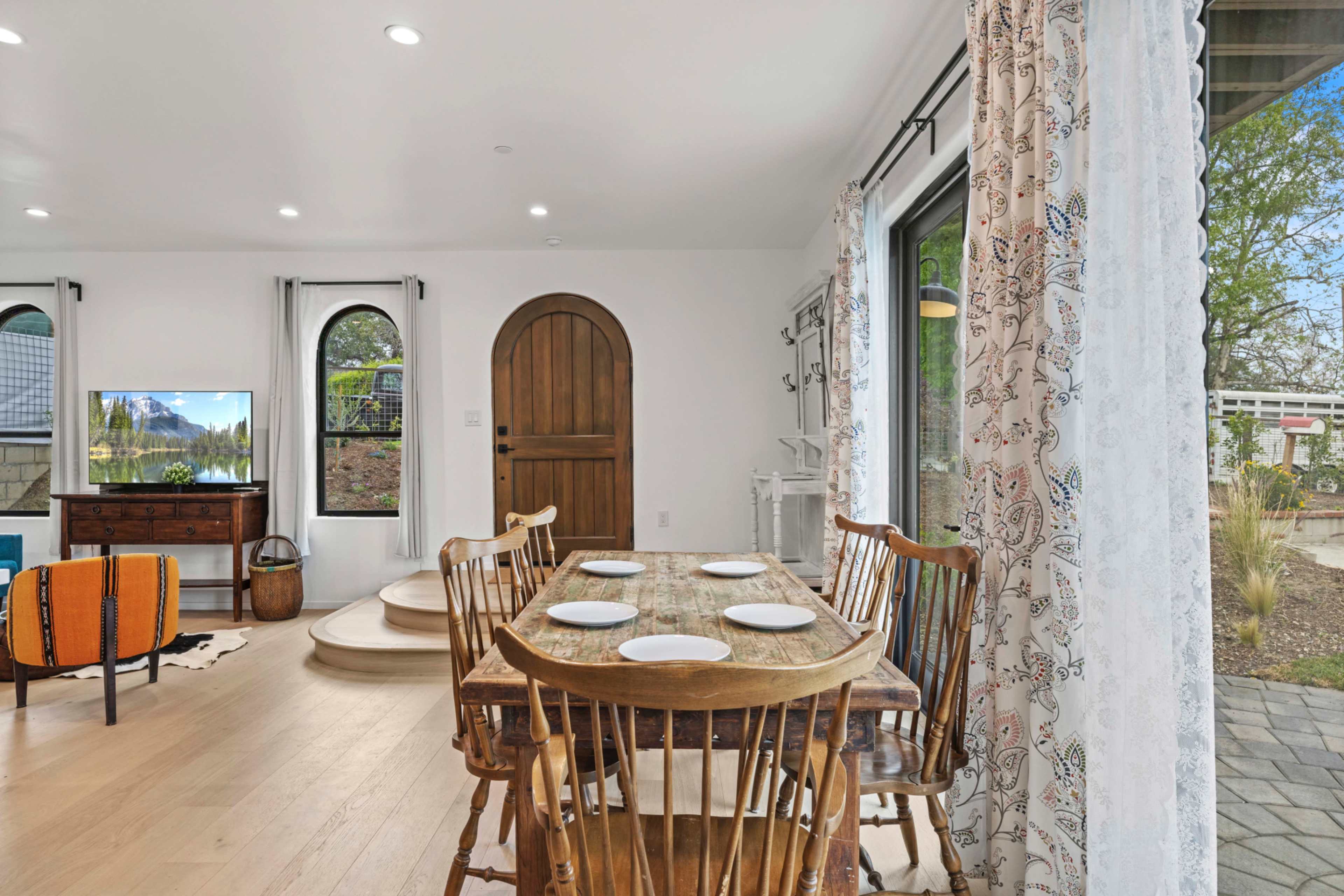 A dining room features a wooden table set for four, surrounded by wooden chairs, with a large window and a doorway leading to an outdoor area.
