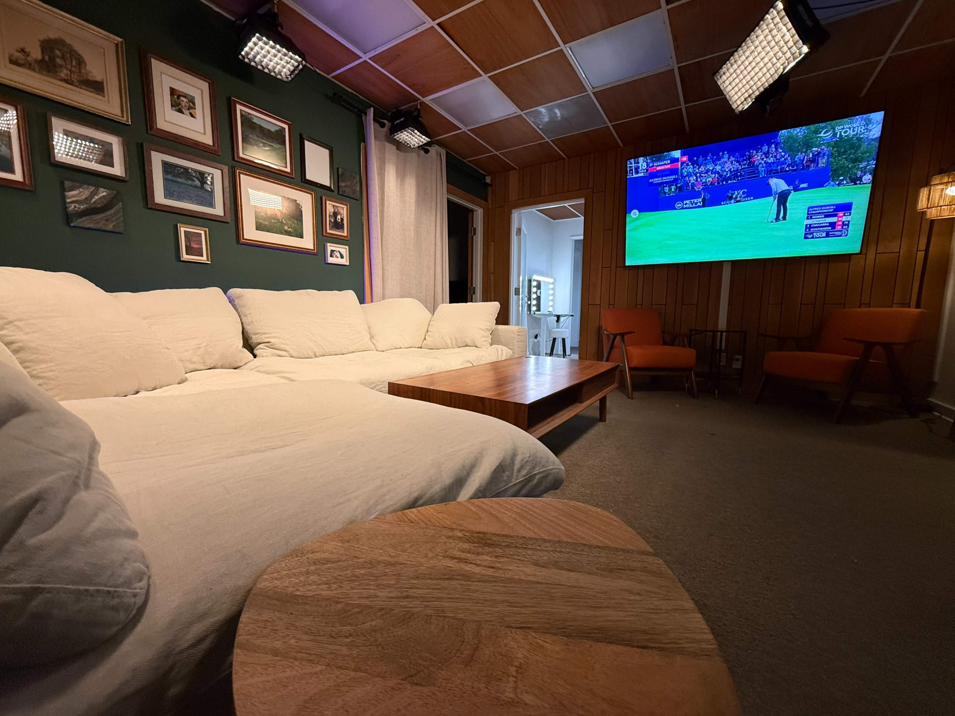 The image shows a cozy living room with a large white sectional sofa, wooden coffee table, and a wall-mounted television displaying a sporting event, surrounded by framed pictures.