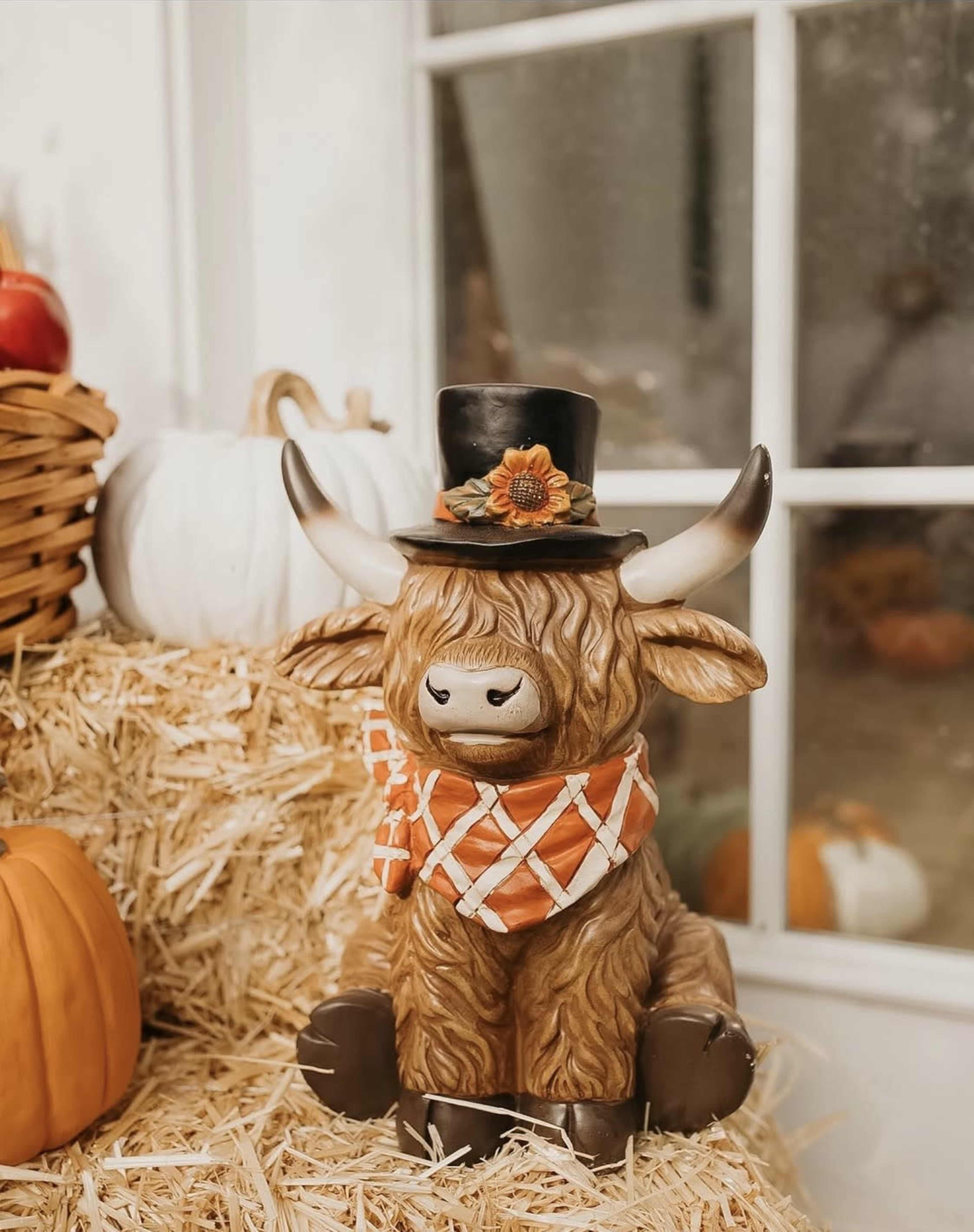A decorative ceramic Highland cow wearing a top hat and scarf sits on a hay bale beside pumpkins and gourds.