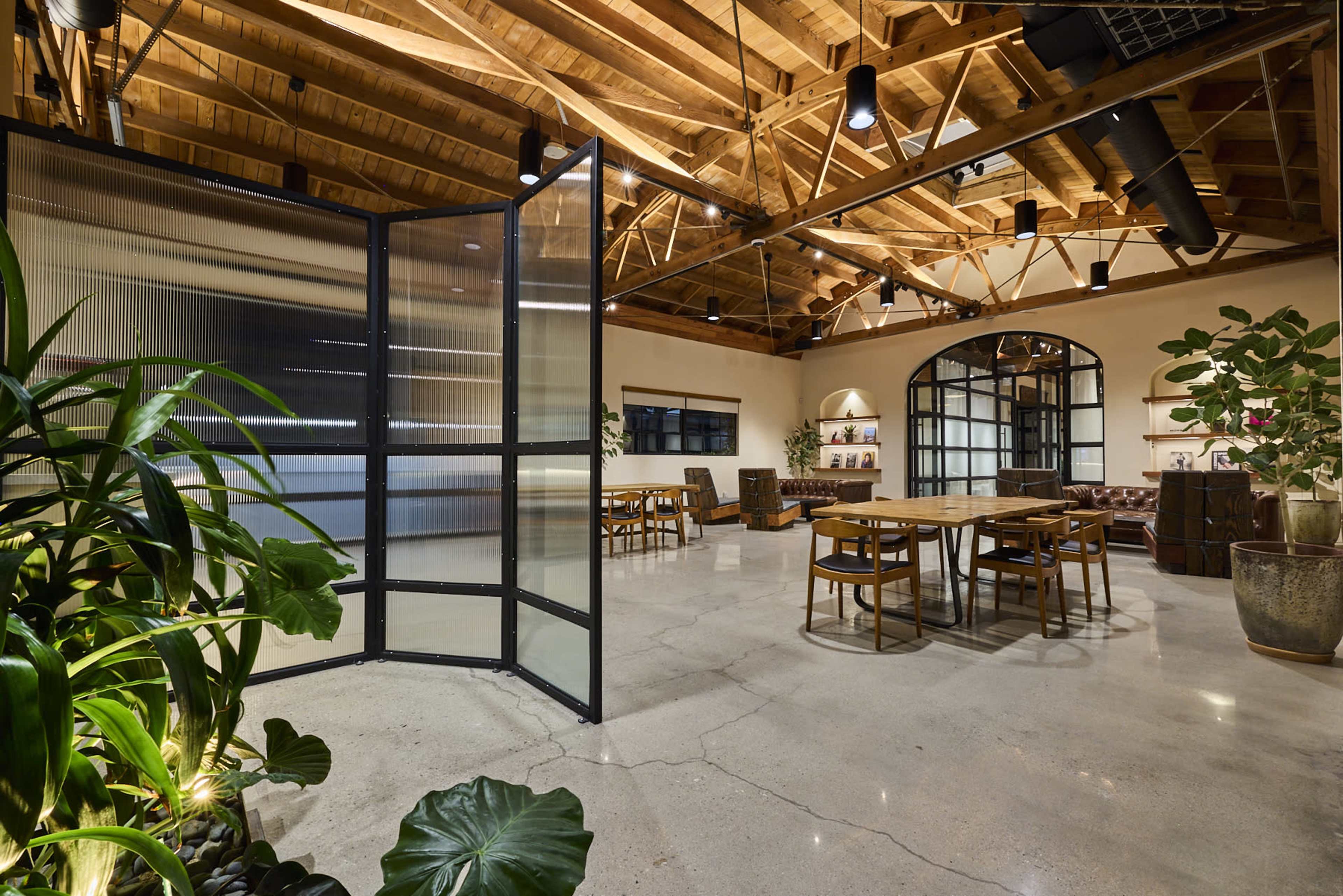 The image shows a spacious, modern workspace featuring wooden beams, a large glass partition, and various seating areas with plants.