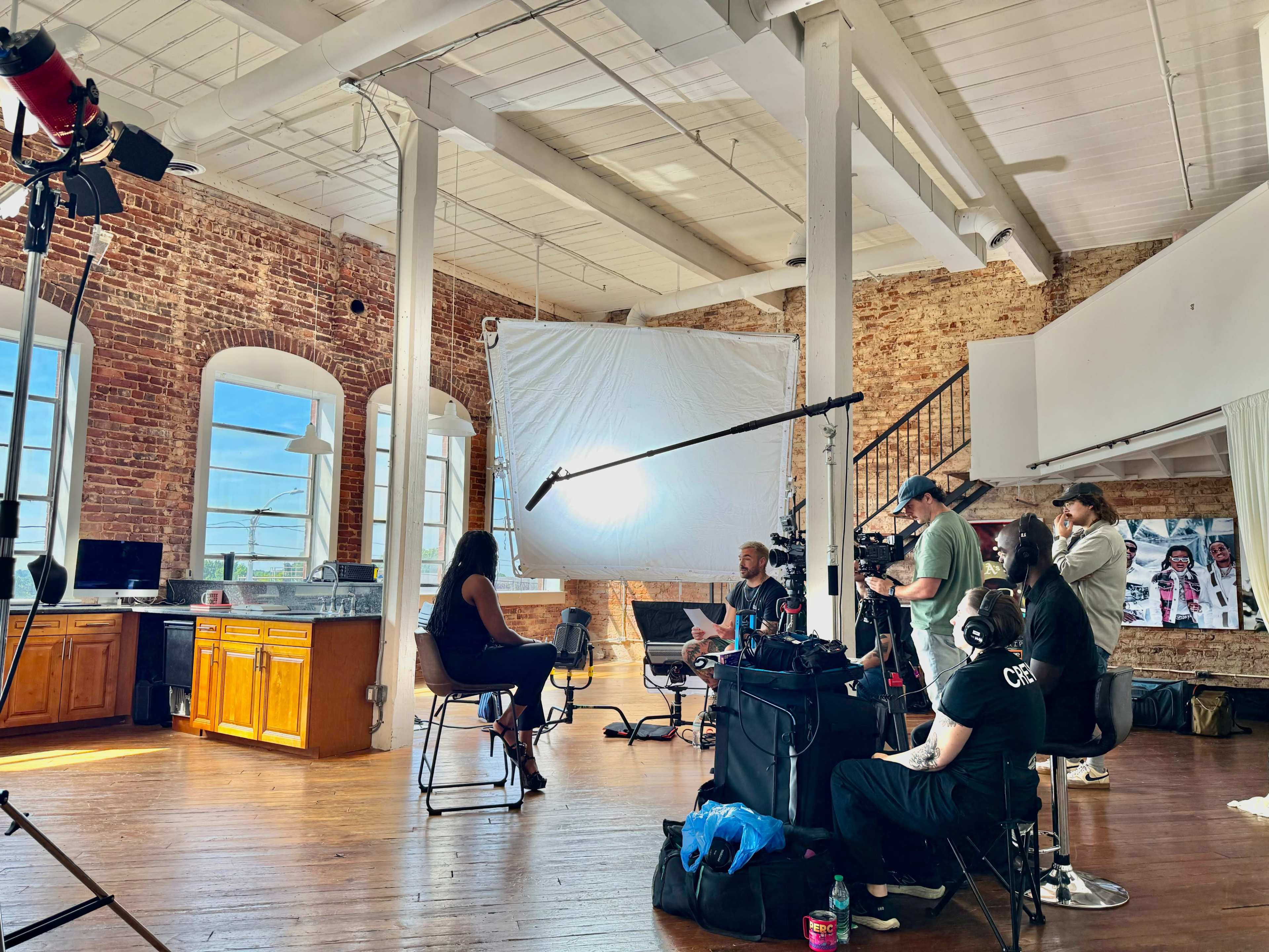 A film crew is set up in a spacious, well-lit studio with exposed brick walls, recording an interview with a seated subject.