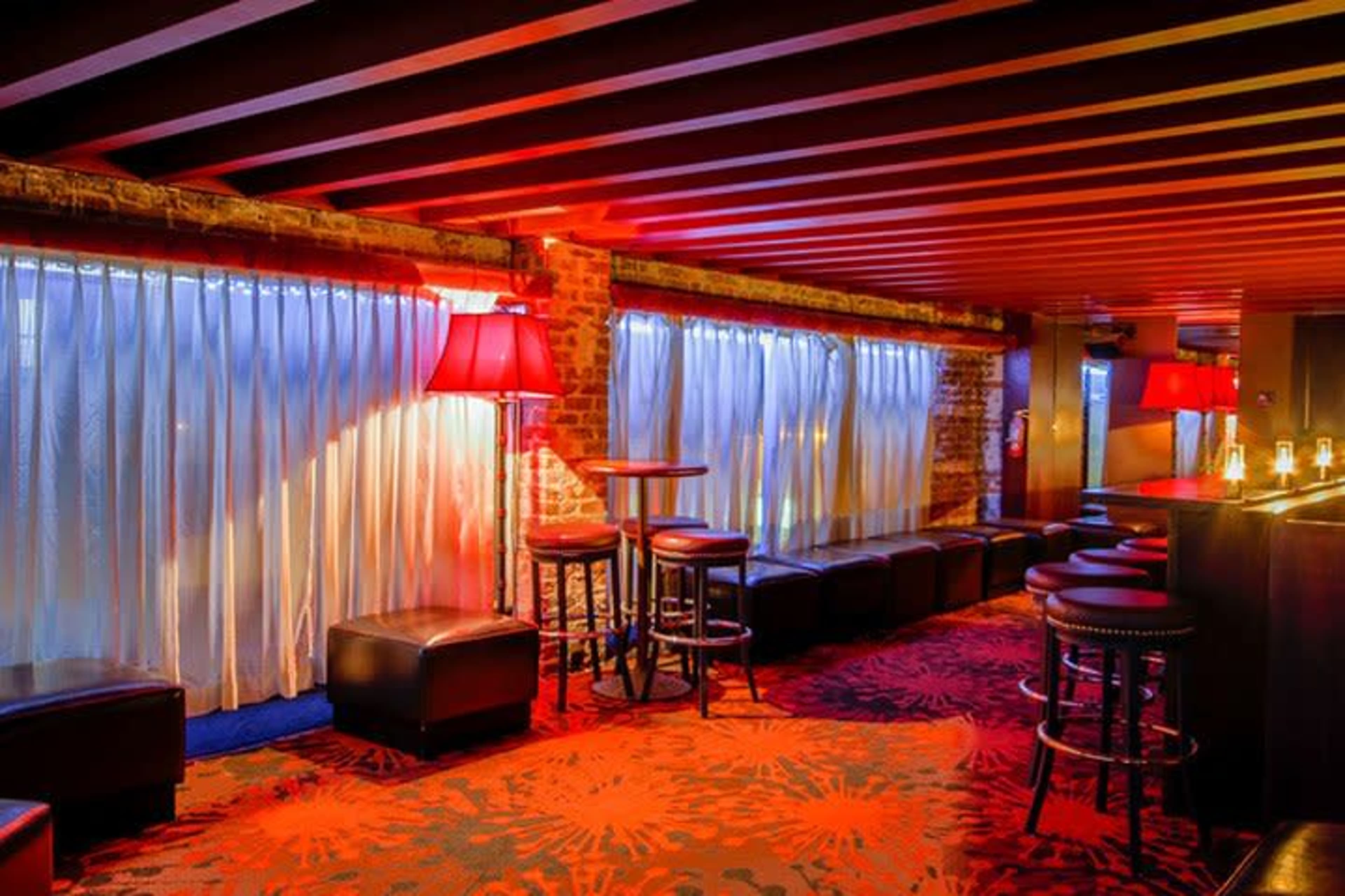 The image shows a dimly lit lounge area featuring red and blue lighting, a patterned carpet, and seating along a wall with curtains.