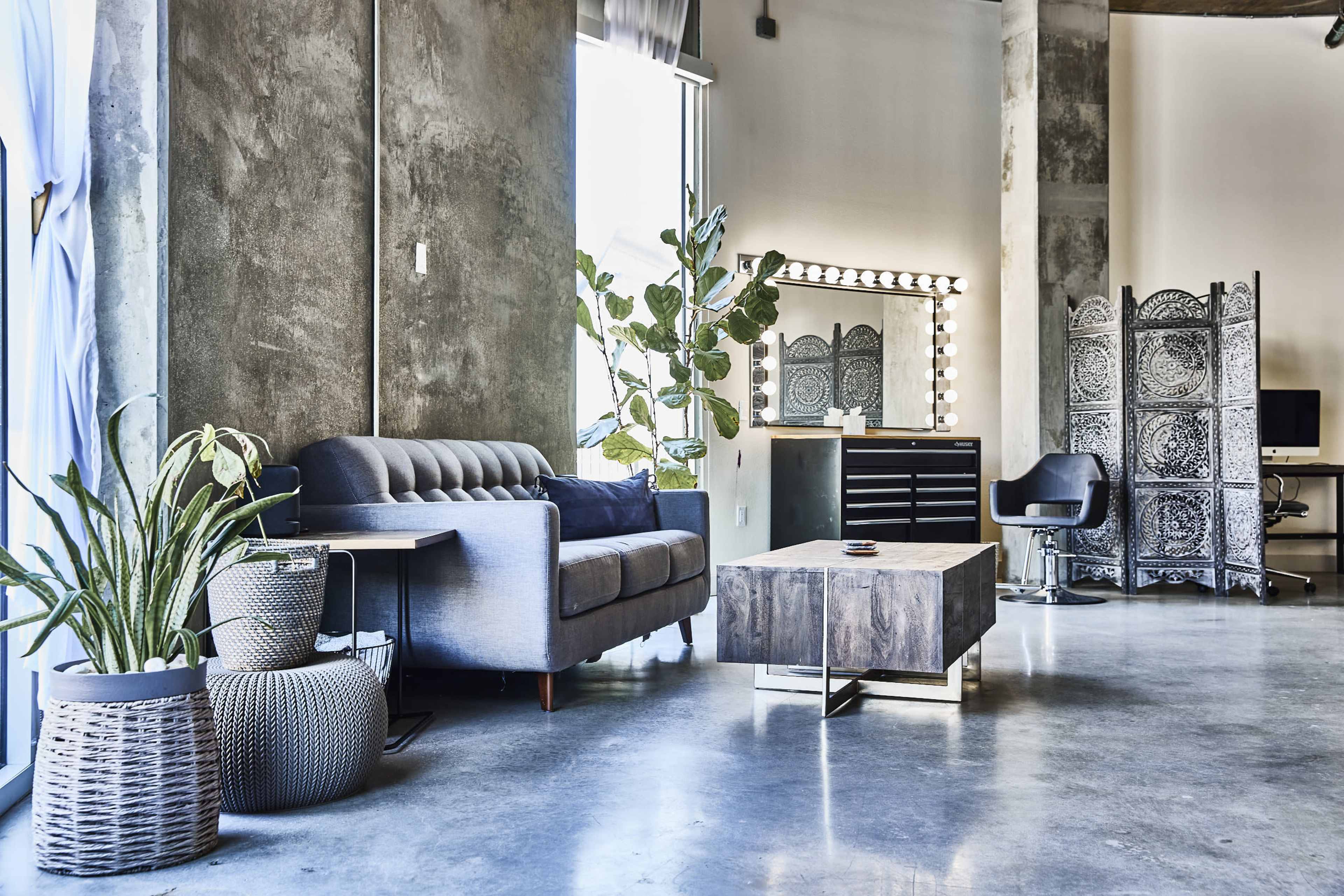 The image shows a modern interior of a stylish salon or lounge, featuring a gray sofa, a wooden coffee table, and a vanity area with bright lights against a concrete wall.