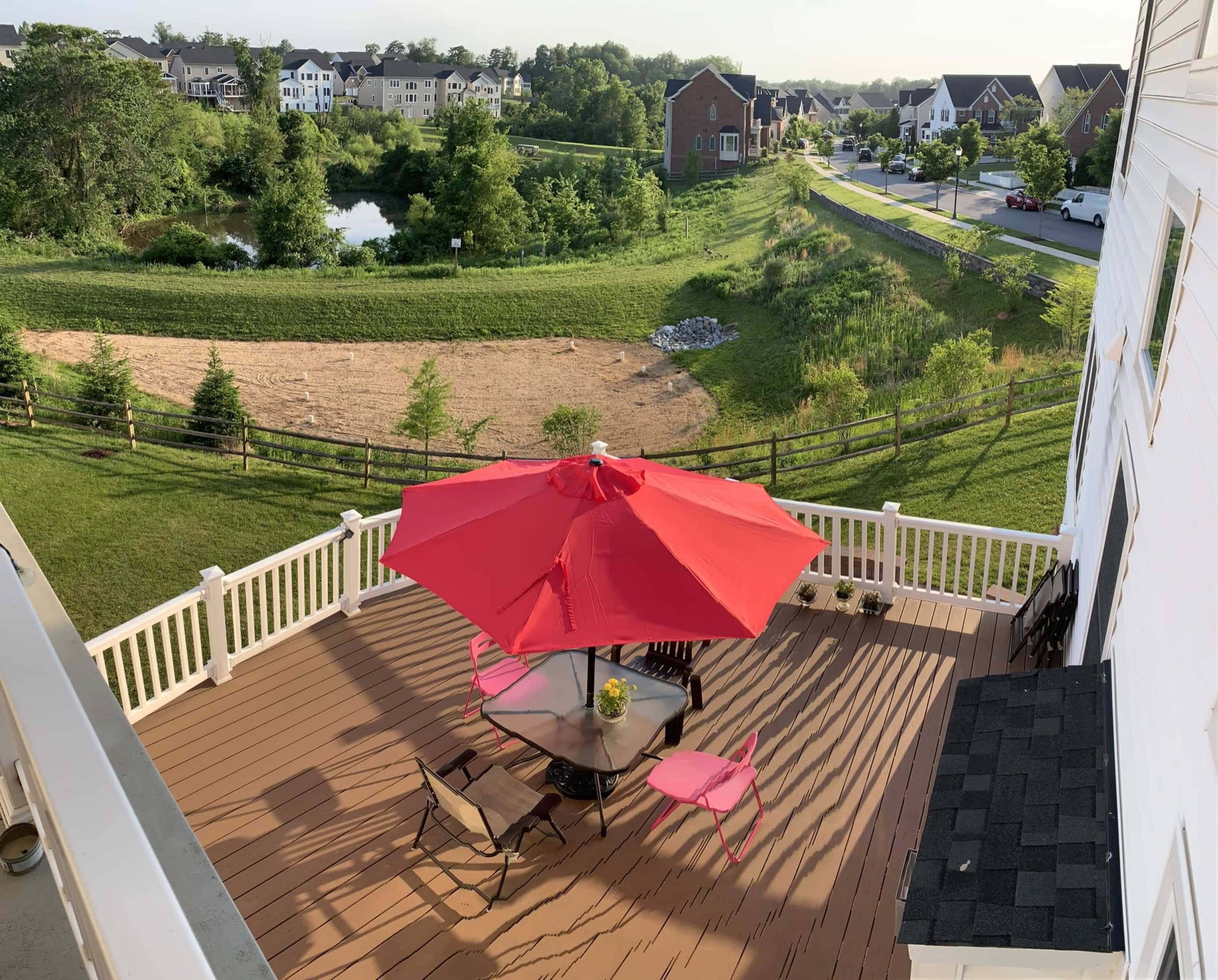 A spacious deck features a round table with black chairs and a large red umbrella, overlooking a grassy area with a pond and nearby houses.