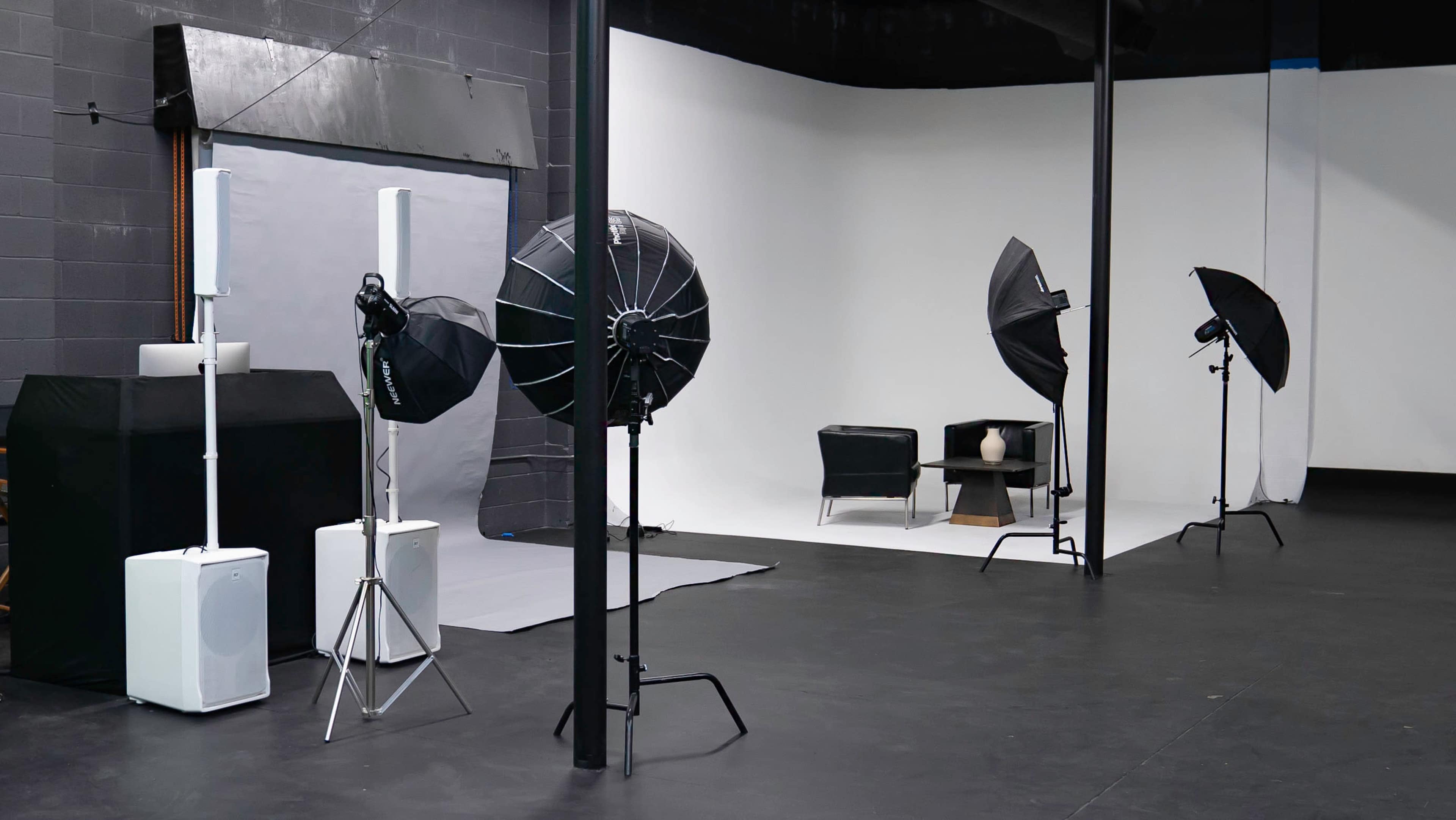 The image depicts a photo studio setup with two softbox lights and a backdrop, featuring a black chair and a table with a vase in the center.