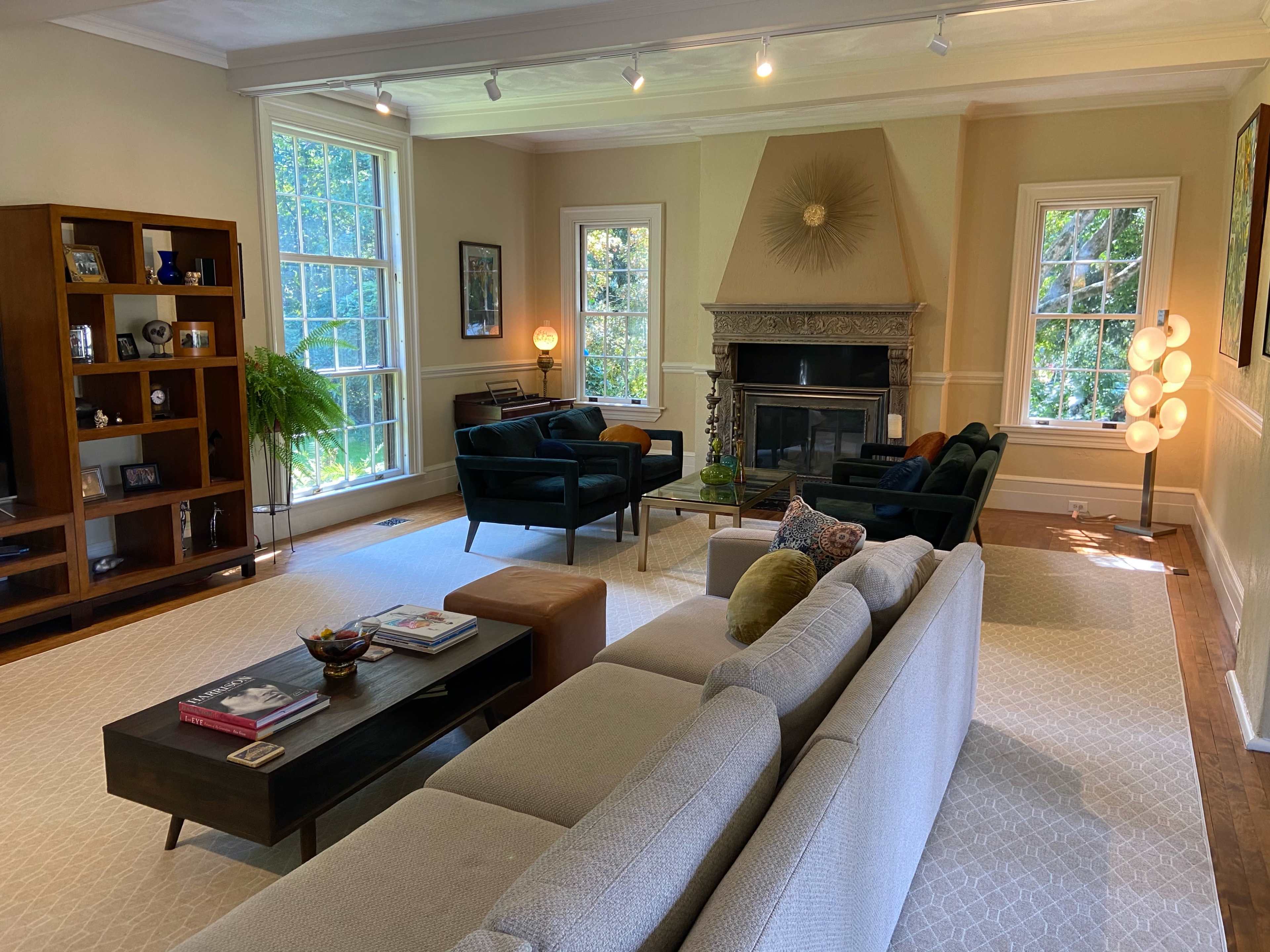 A living room with a light-colored sectional sofa, a coffee table, a fireplace with ornate detailing, and large windows allowing natural light to enter.