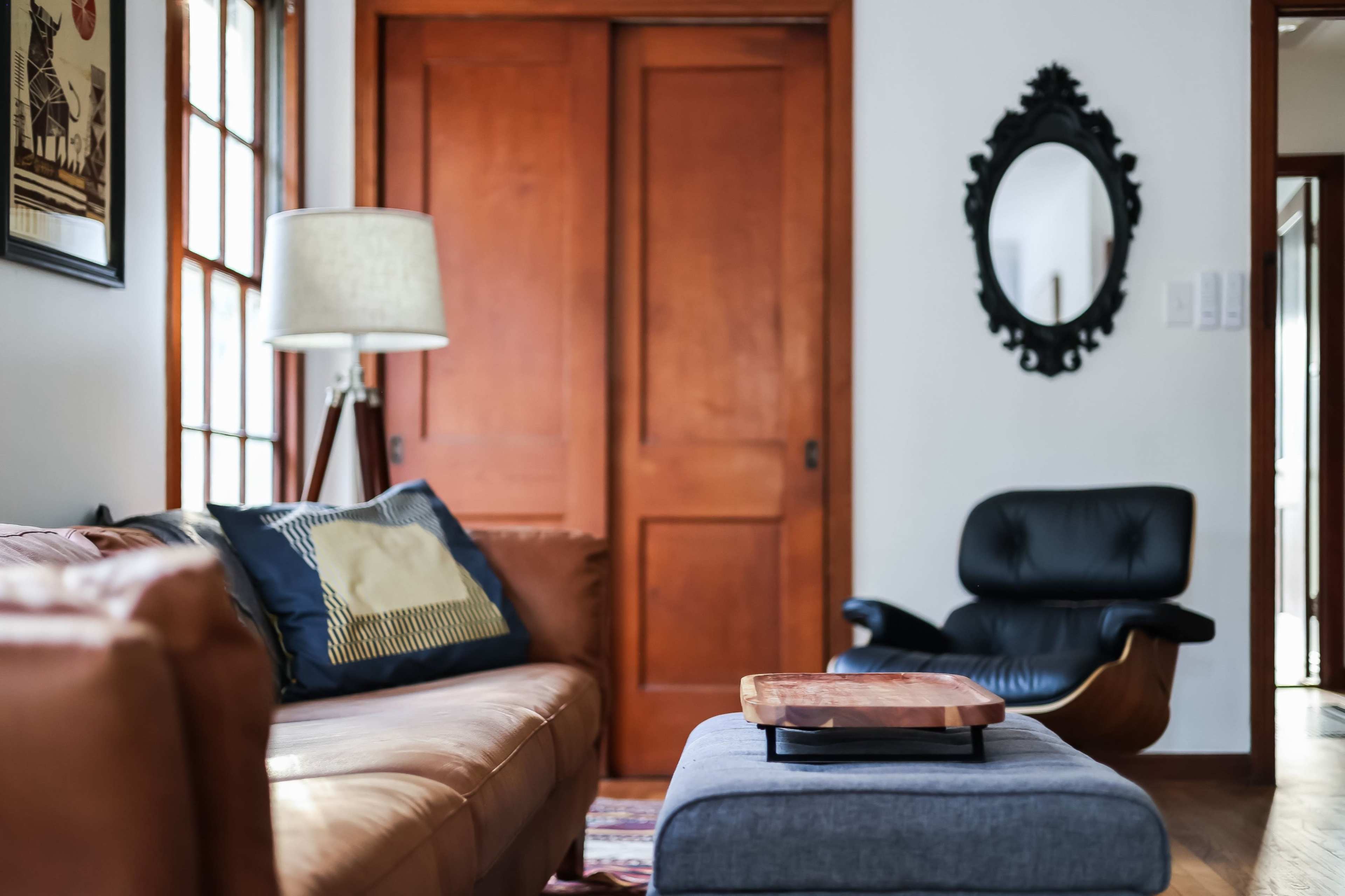 The image shows a living room with a brown leather sofa, a blue ottoman with a wooden tray on top, a black lounge chair, and a floor lamp beside a wall with wooden doors.