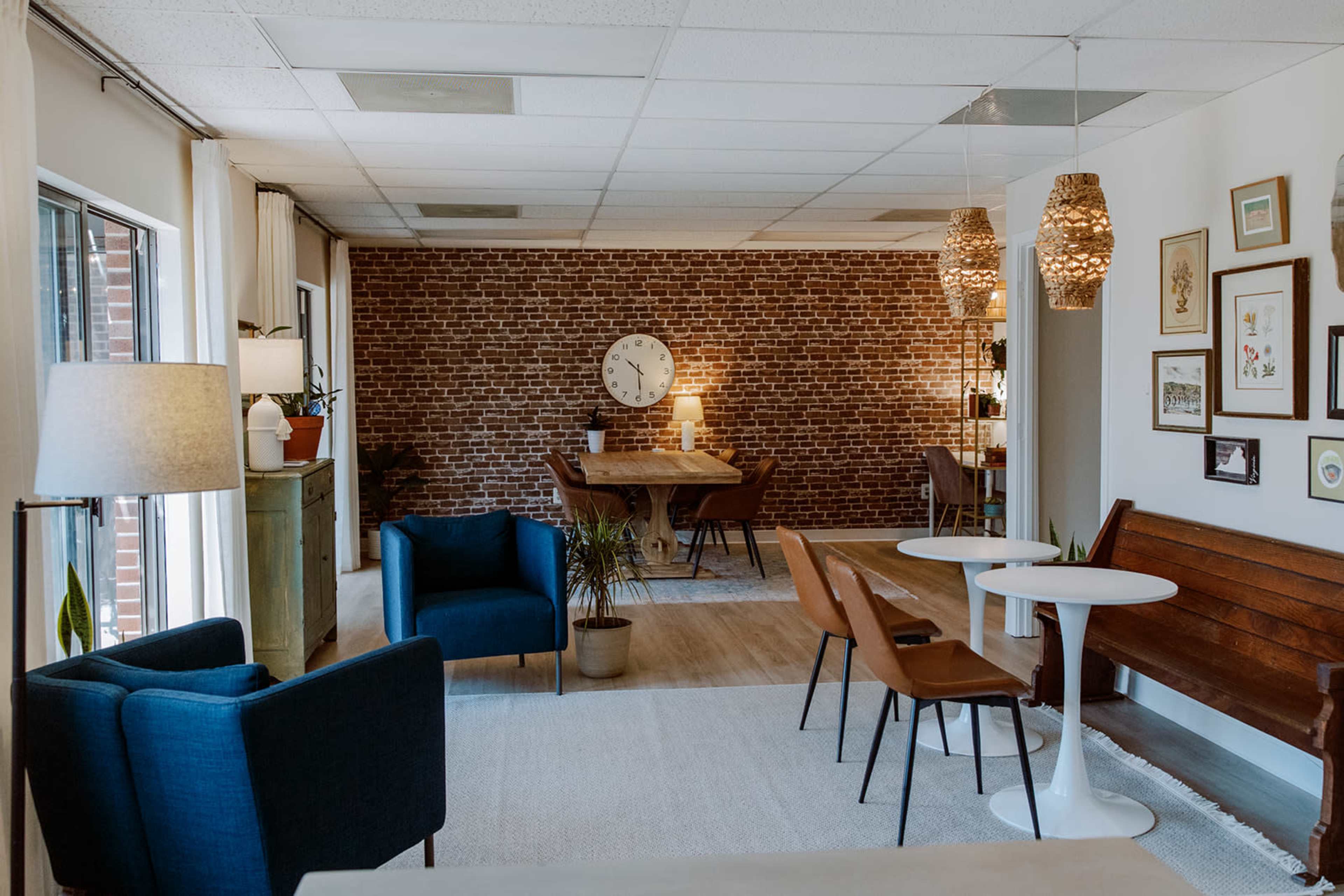 The image depicts a modern, open space featuring a combination of seating areas, a wooden dining table, and a brick accent wall with decorative lighting and artwork.