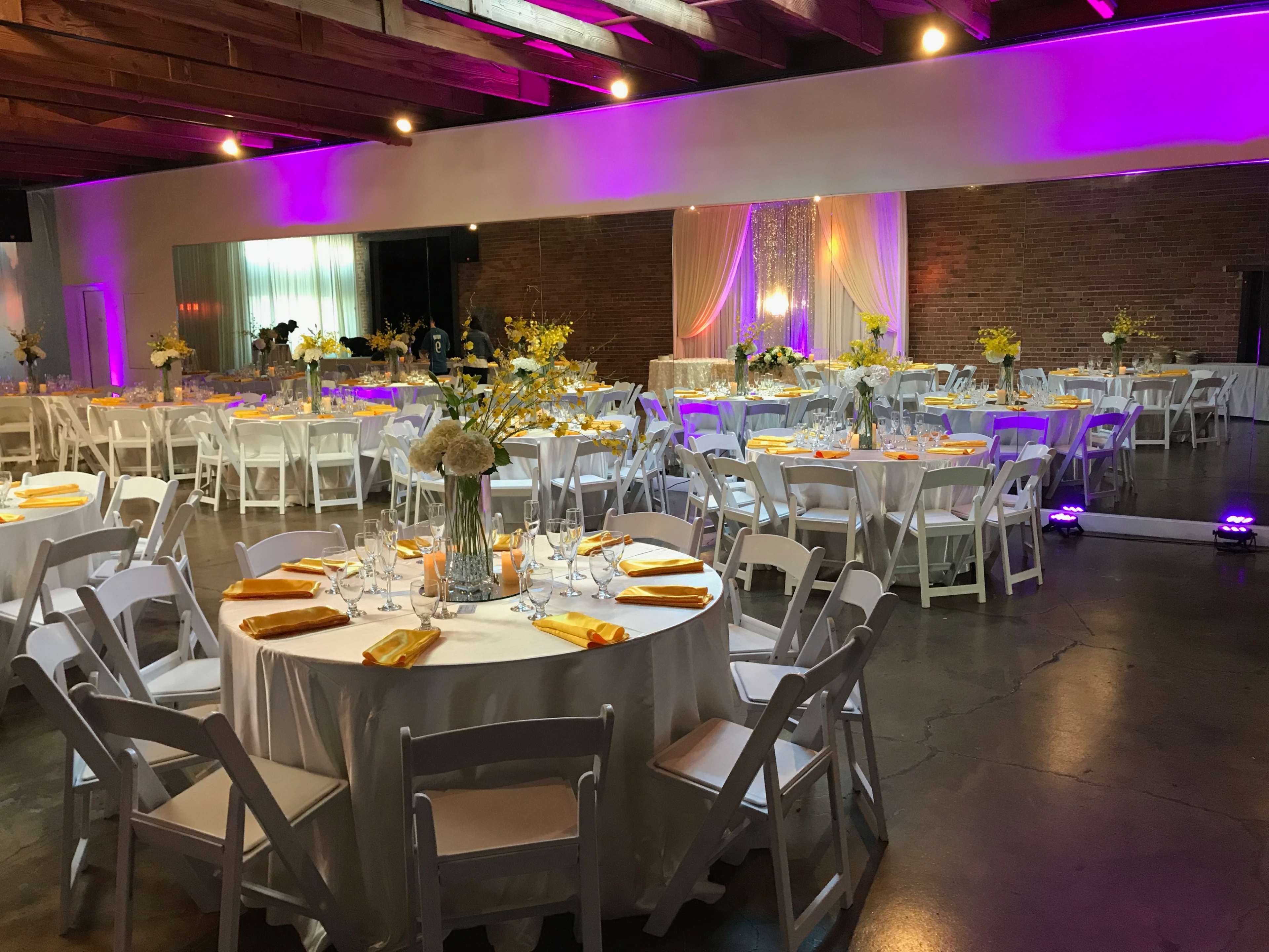 A wedding reception venue features round tables set with yellow napkins and floral centerpieces under colorful lighting with a backdrop of draped fabric.