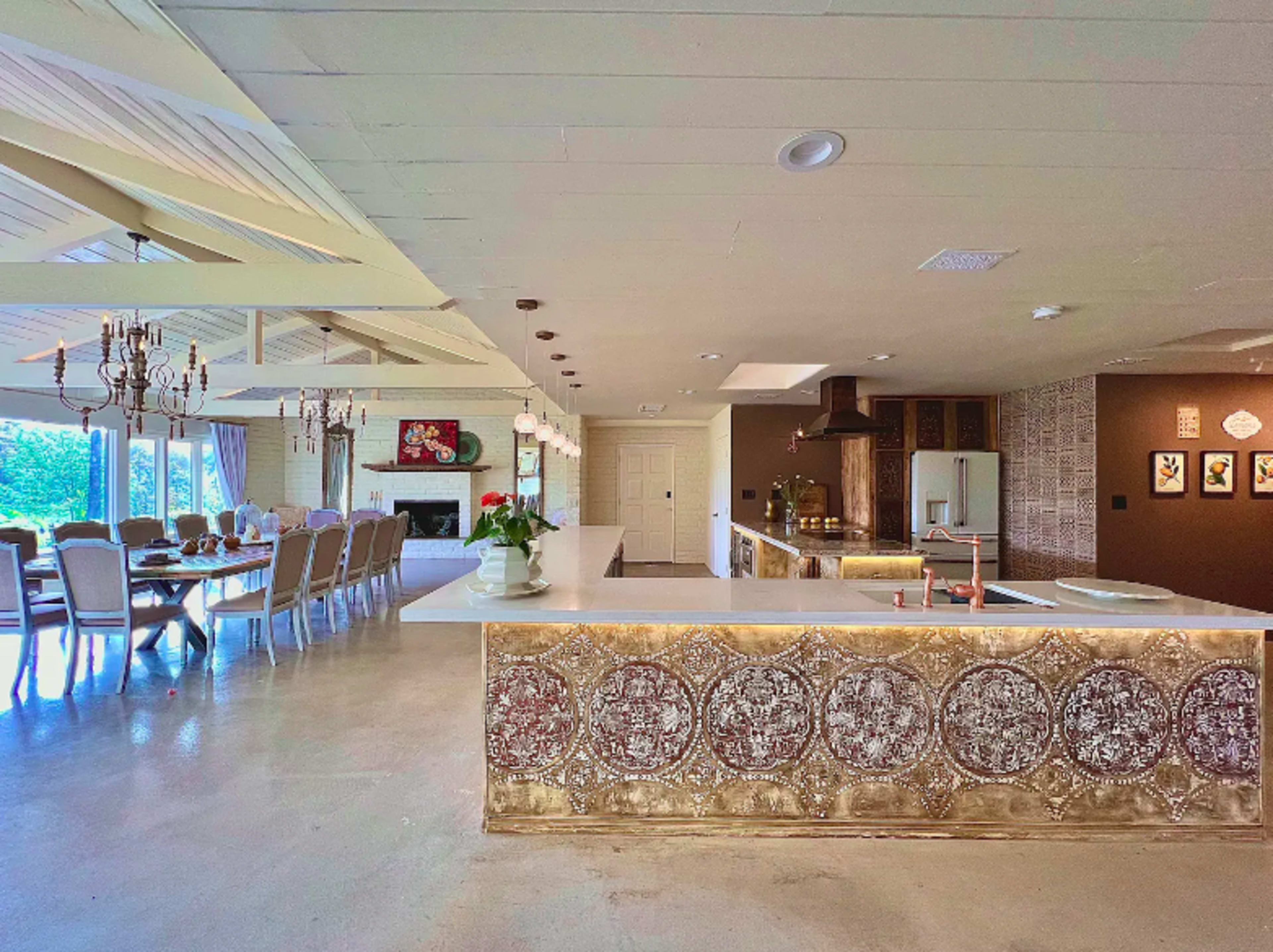 The image shows a modern kitchen and dining area with a large white countertop, elegant dining chairs, and a decorative wall feature.