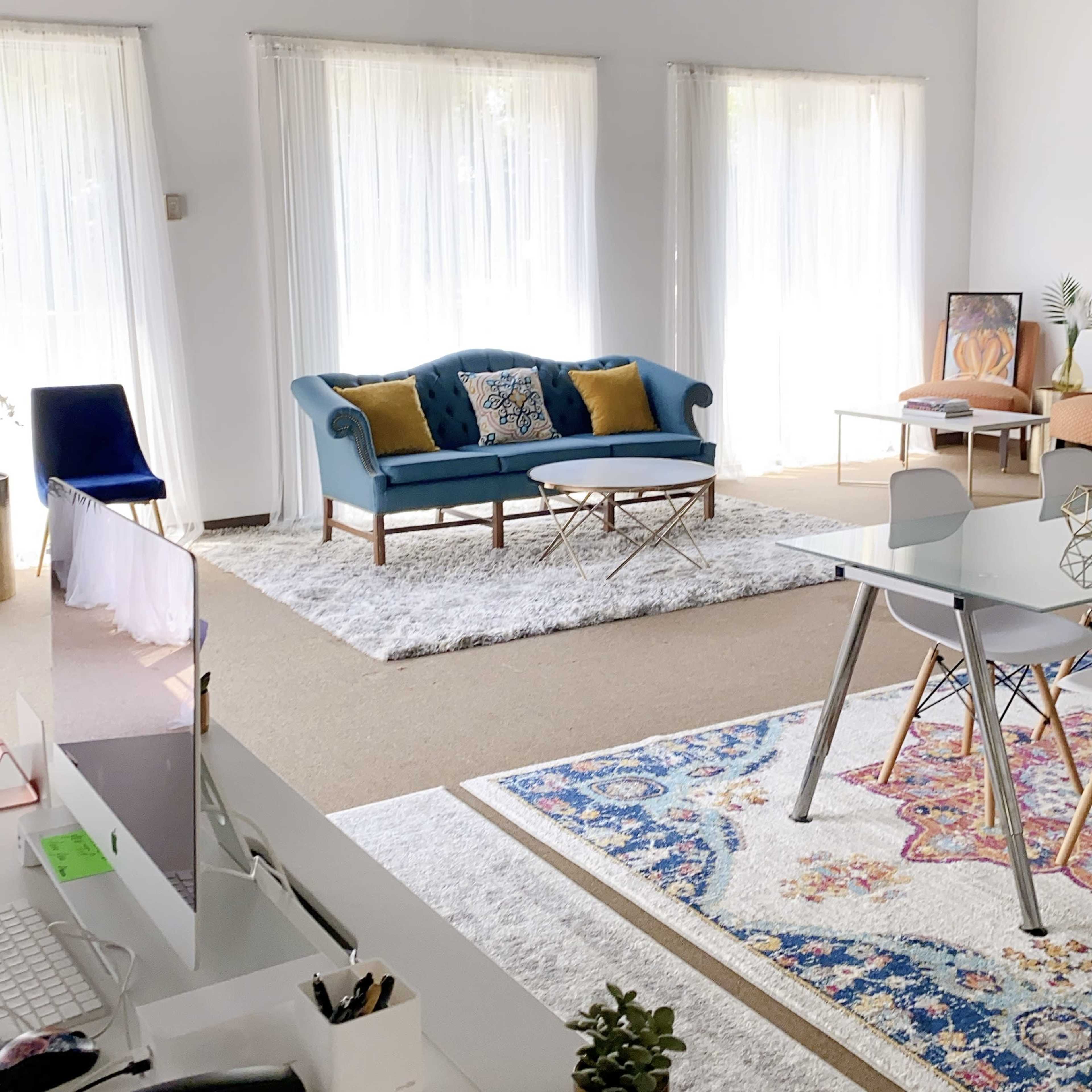 A bright room features a blue sofa with yellow cushions, a glass coffee table on a white rug, and a dining area with a patterned rug and modern chairs.