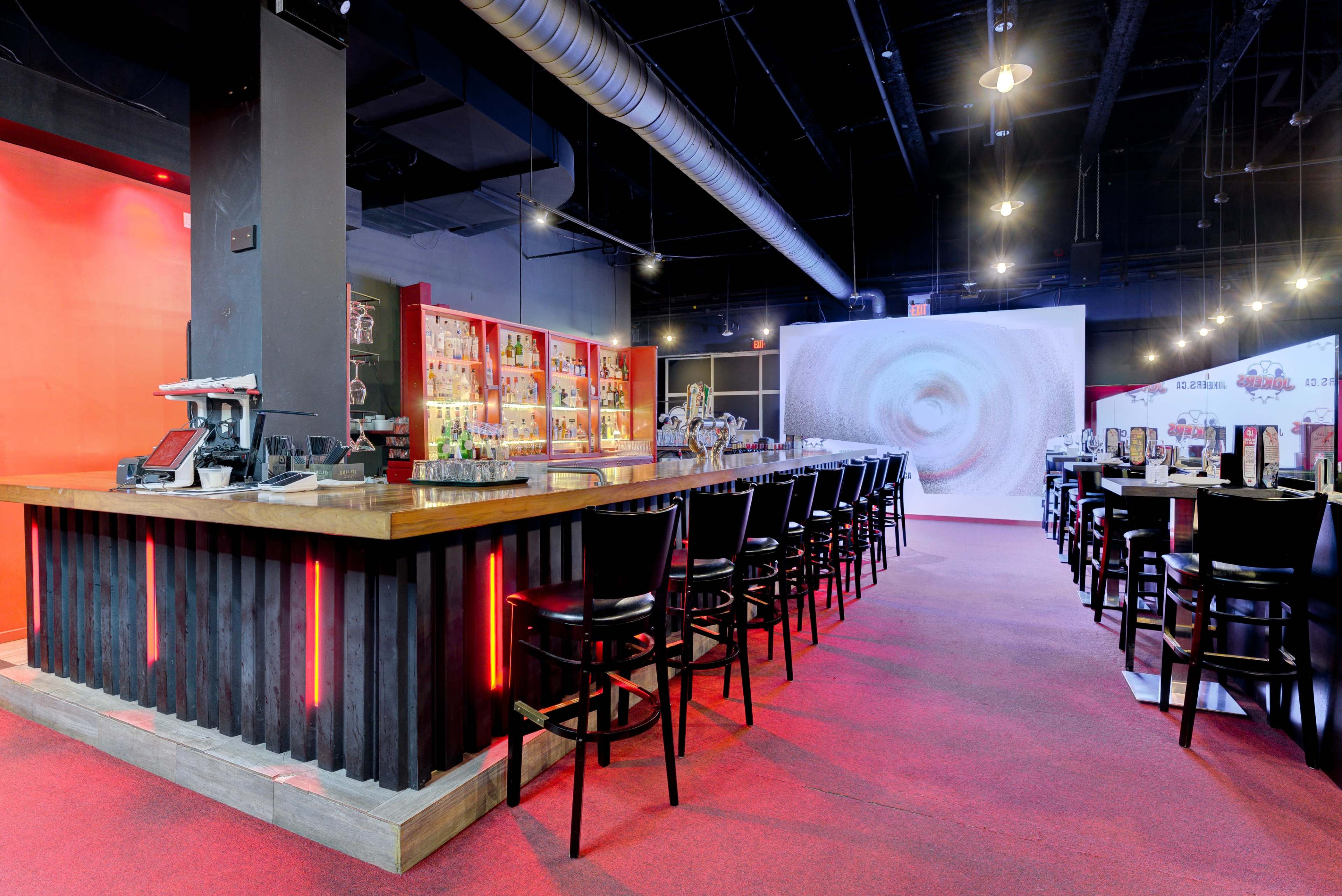 A modern bar area with a long wooden counter, high chairs, and illuminated shelves behind the bar, featuring various bottles.