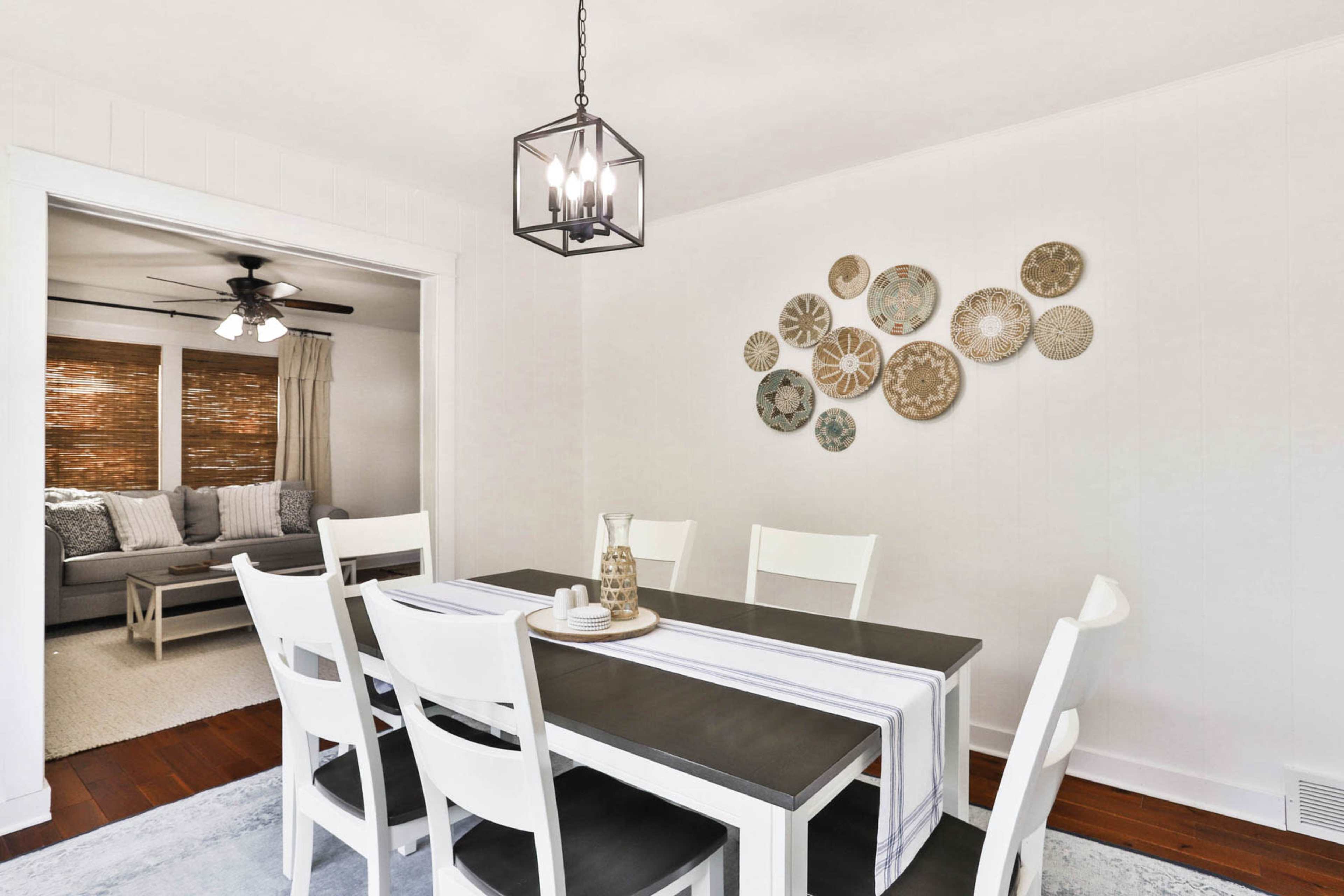 A dining room features a rectangular table with white chairs, centered under a black lantern light, and a wall adorned with decorative plates.