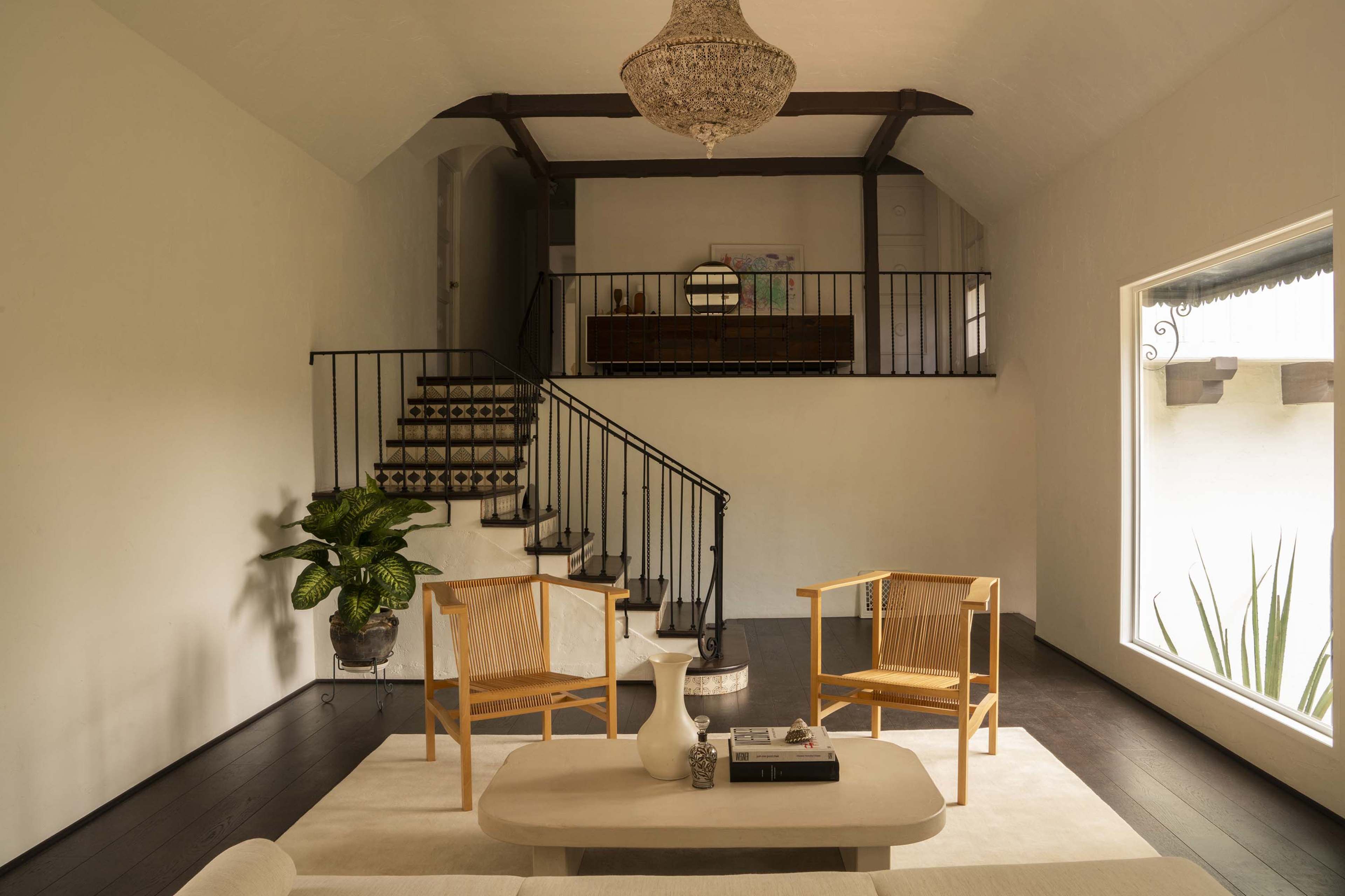 A spacious living area features a staircase leading to a loft, with two wooden chairs and a coffee table on a light rug beneath a chandelier.