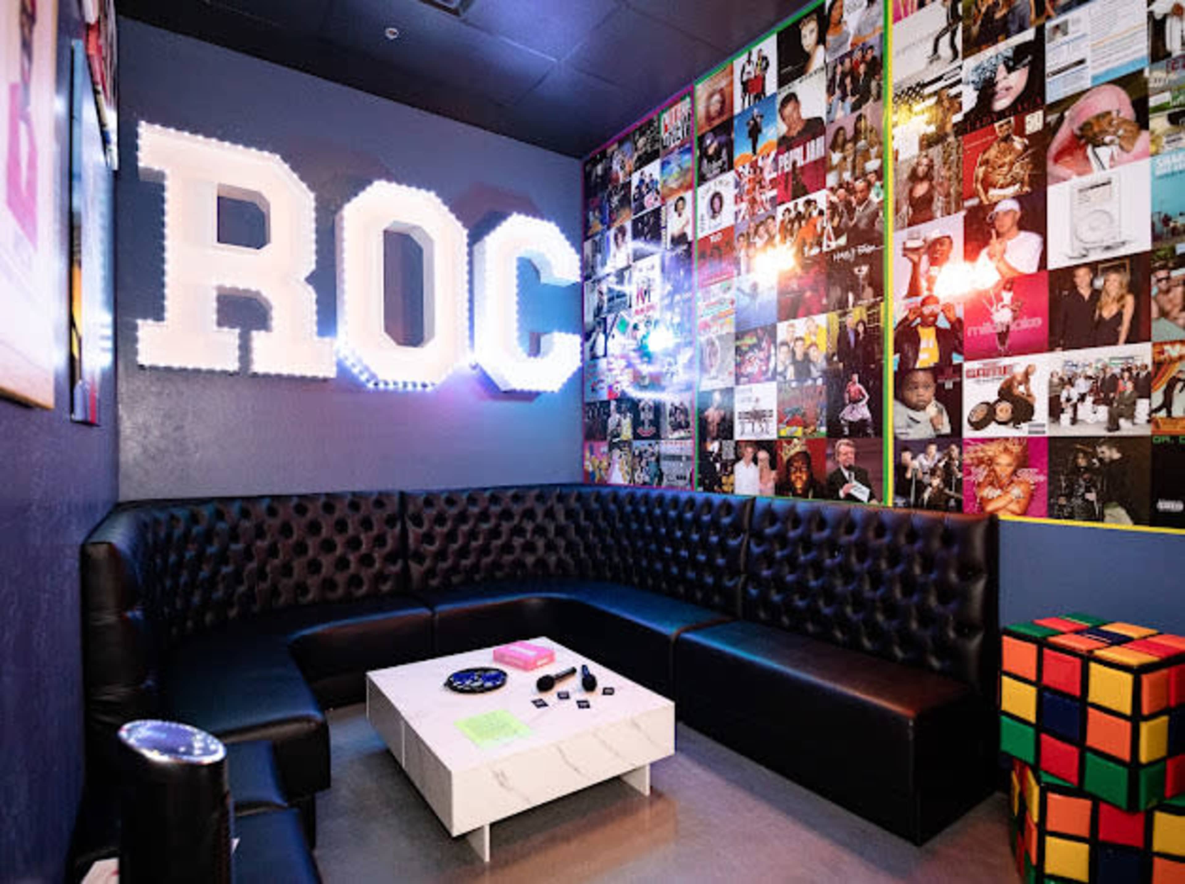 A small lounge area with a black leather sectional sofa, a white coffee table, and a wall adorned with colorful album covers.
