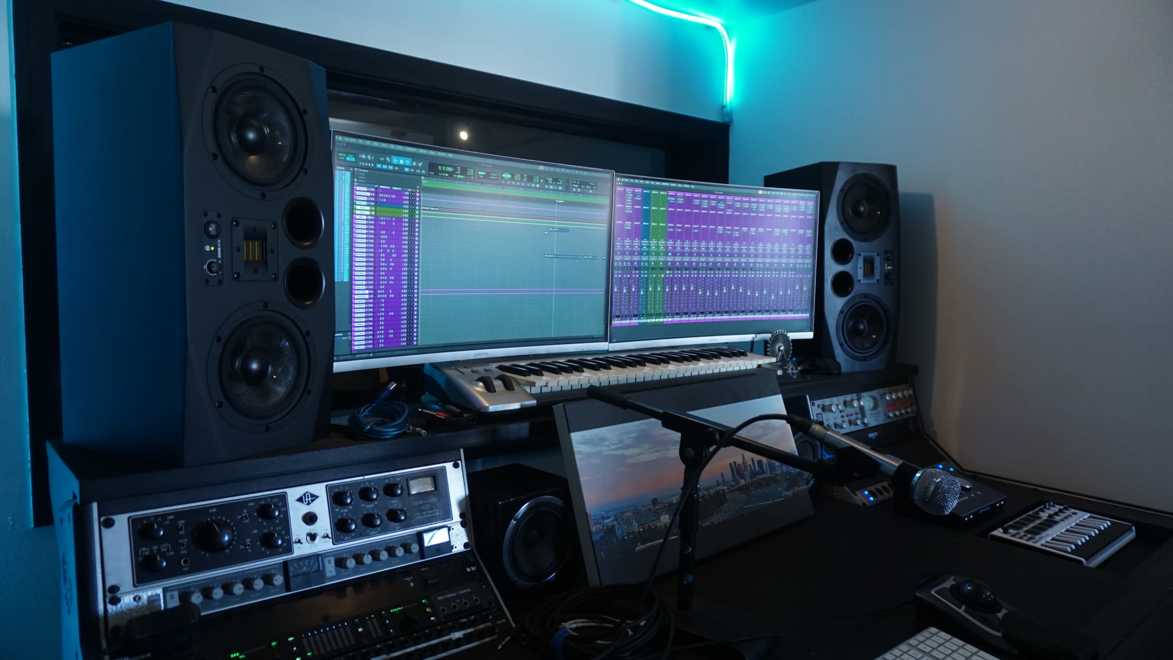 The image shows a music production setup featuring two large studio monitors, a keyboard, multiple audio interface units, and two computer screens displaying a digital audio workstation.