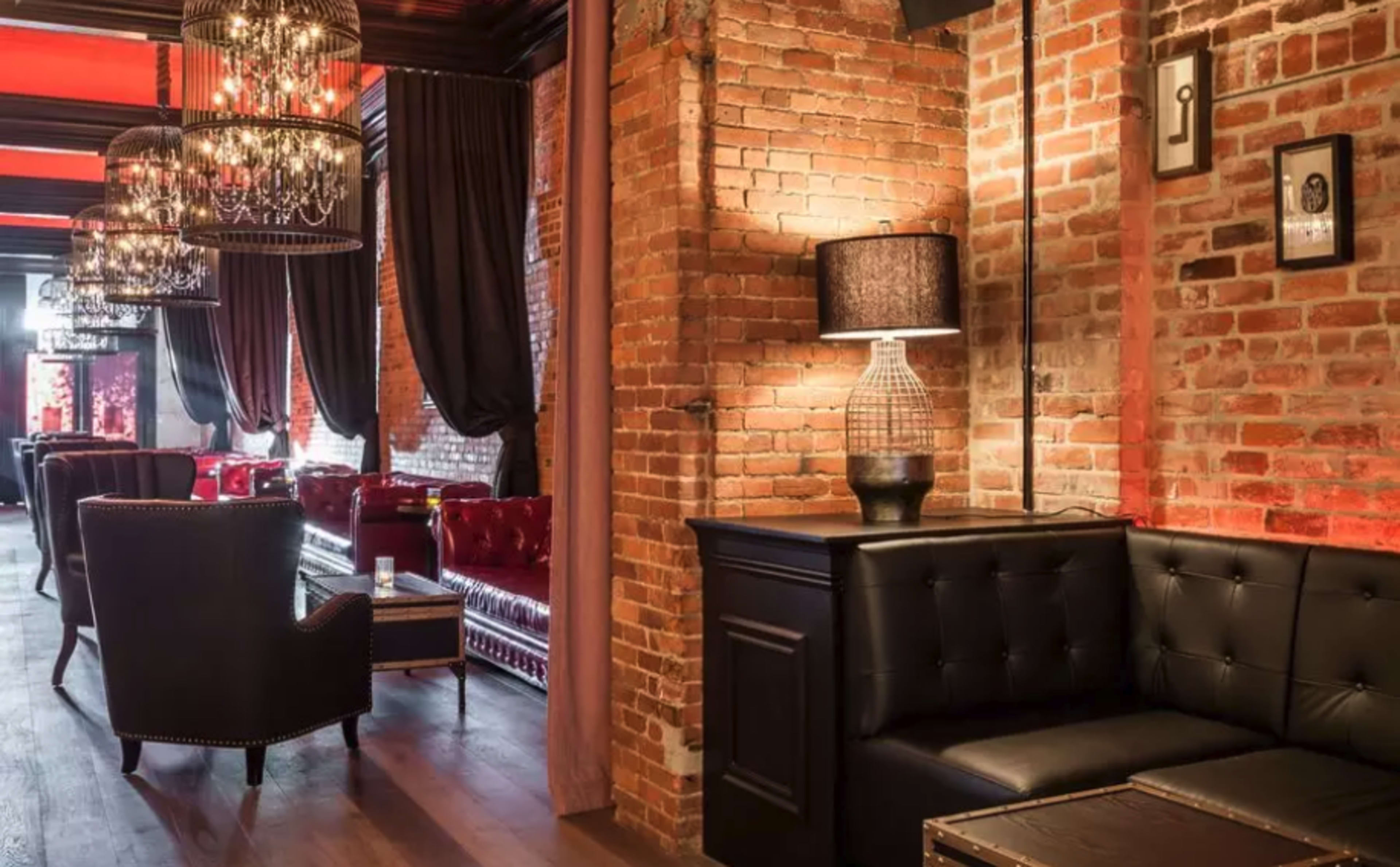 The image shows a stylish lounge with exposed brick walls, plush seating, and ornate chandeliers.