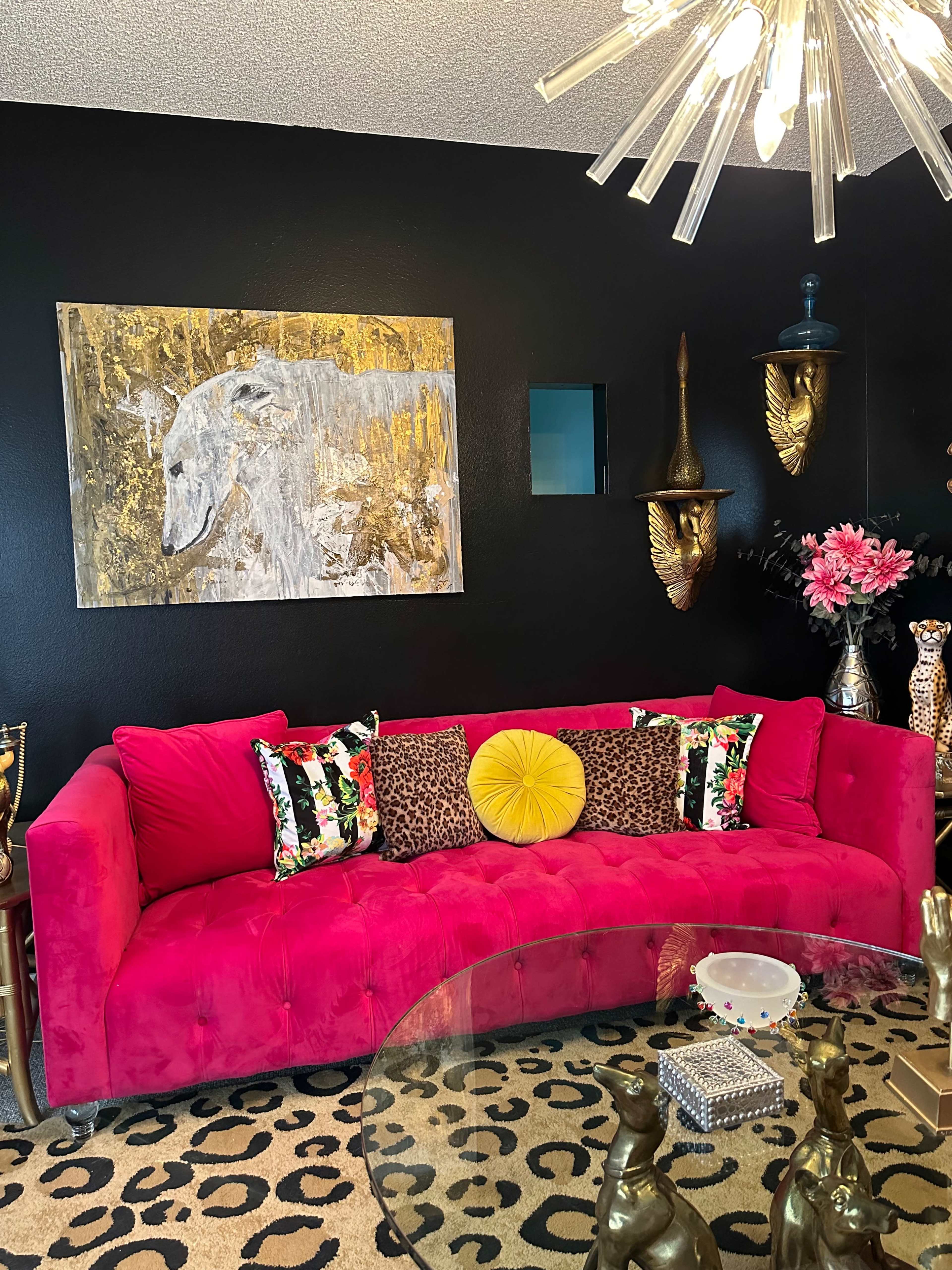 A vibrant pink sofa with patterned cushions in a room featuring a black wall, a colorful painting, and decorative accents on shelves.
