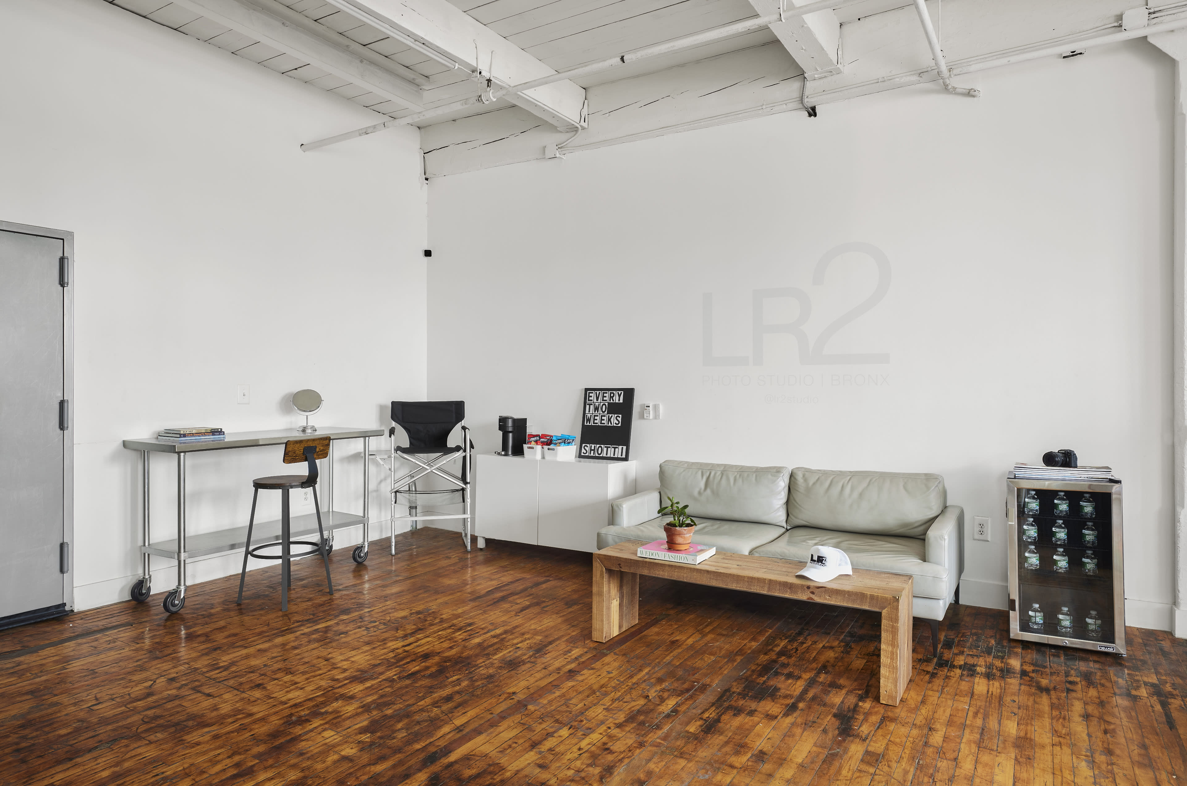 A minimalist studio space with a wooden floor, a green sofa, a coffee table, a desk with a stool, and a small refrigerator.