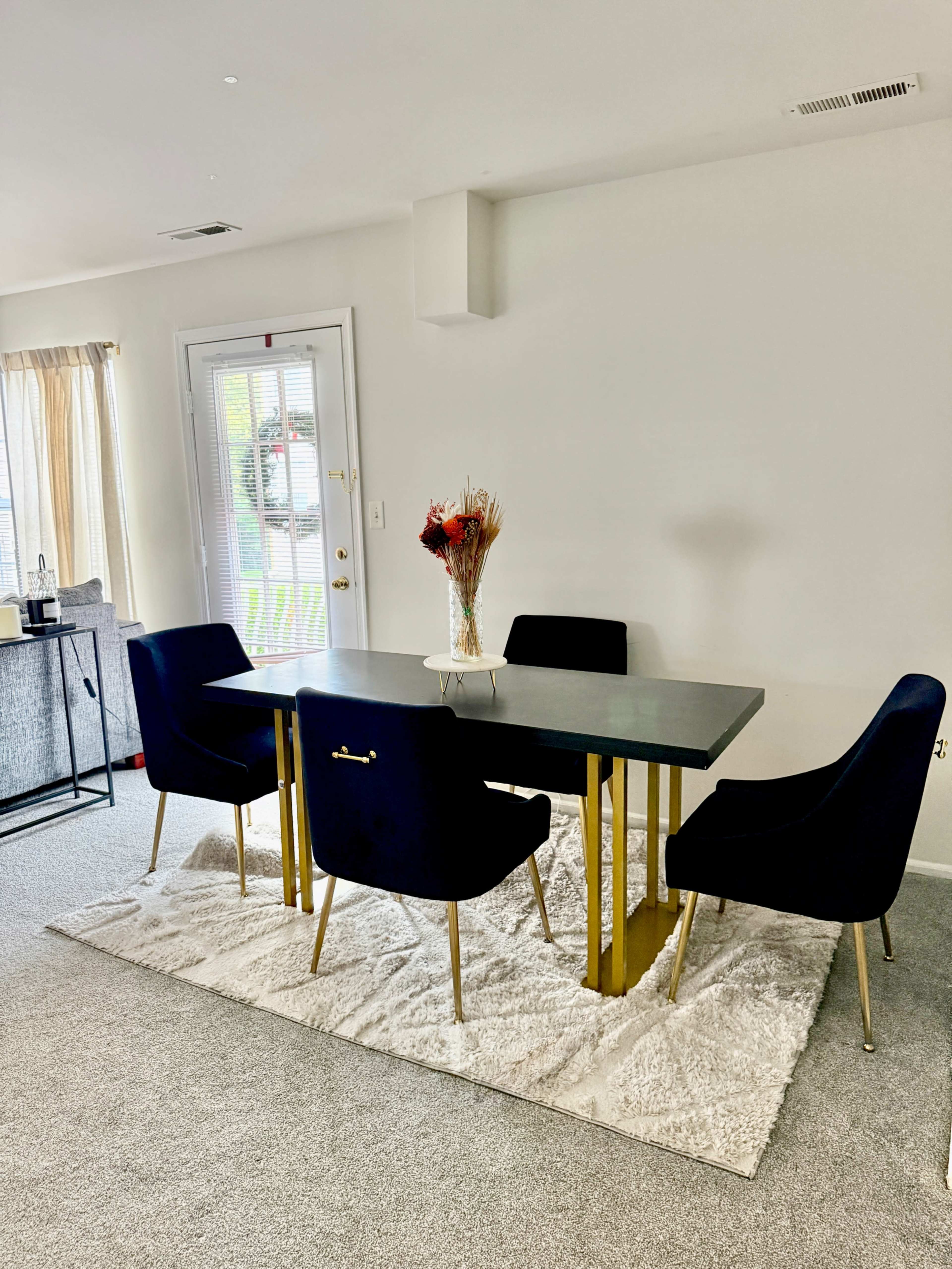 A modern dining area features a black table with gold legs surrounded by four dark blue chairs, all set on a light-colored rug.