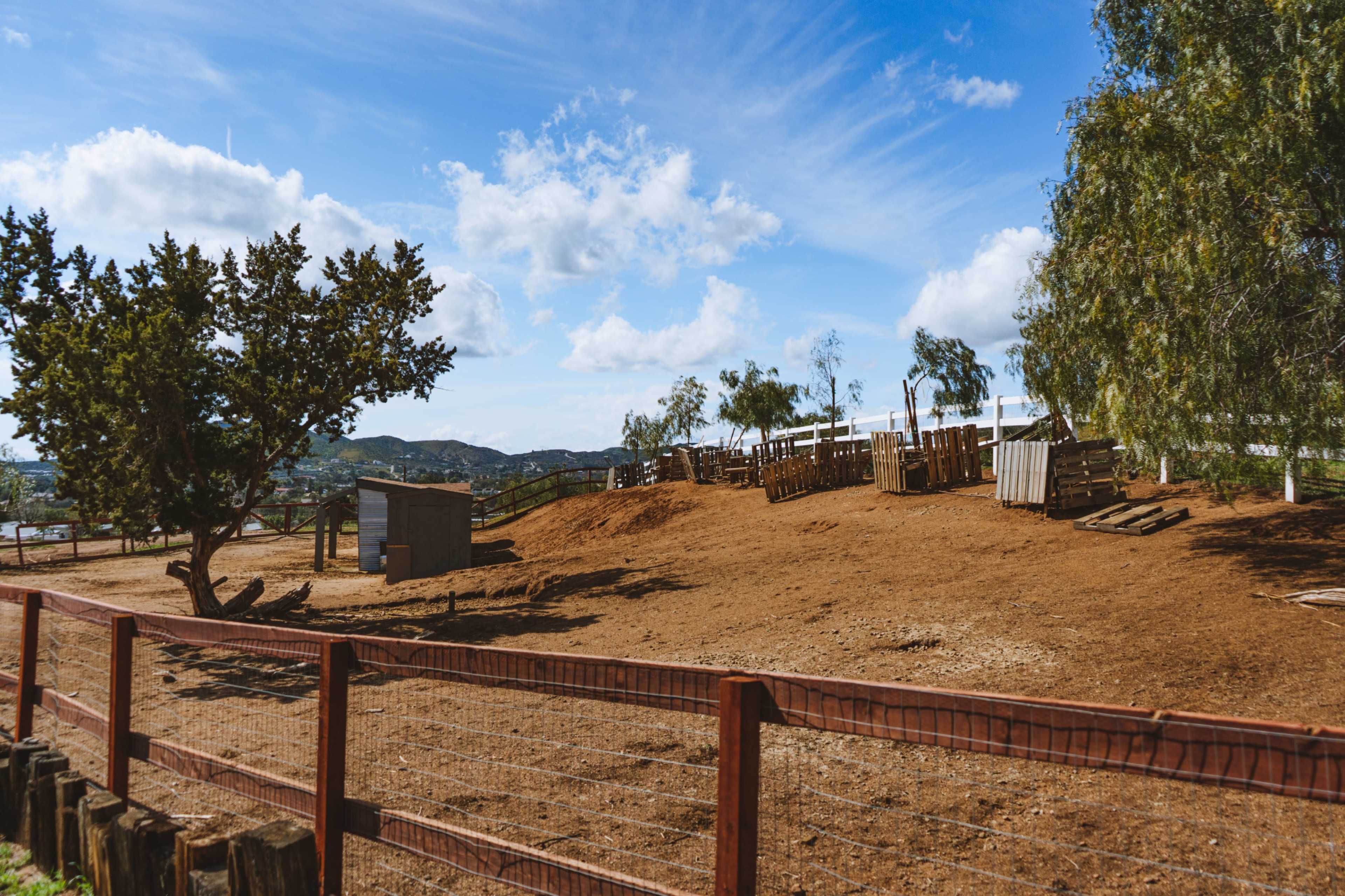 The image shows a fenced dirt area on a farm with a tree, a shed, and wooden structures in the background under a blue sky.