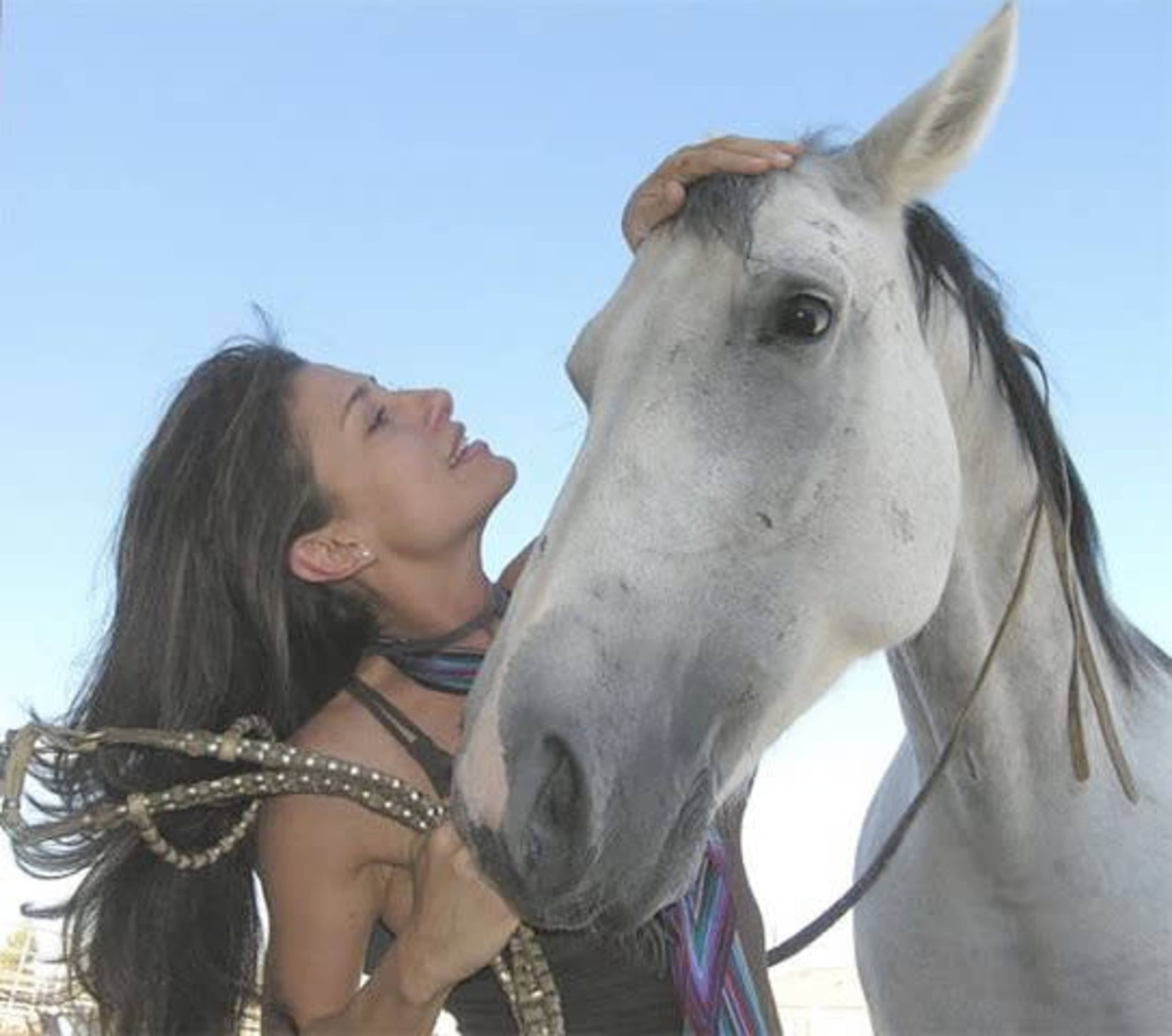 A woman affectionately interacts with a gray horse against a clear blue sky.
