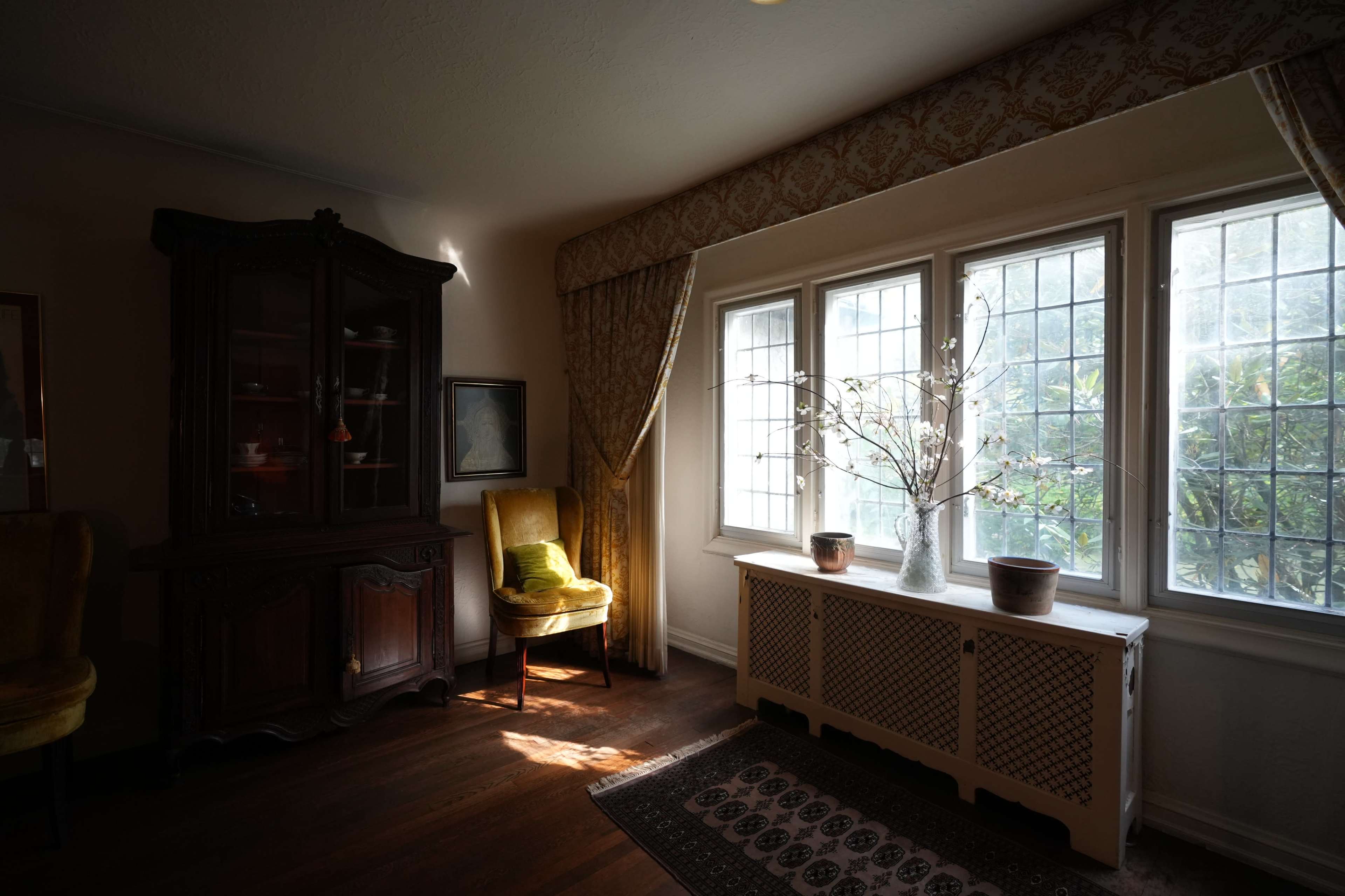 The room features a wooden cabinet, a chair, and a large window with natural light casting soft shadows on the floor.