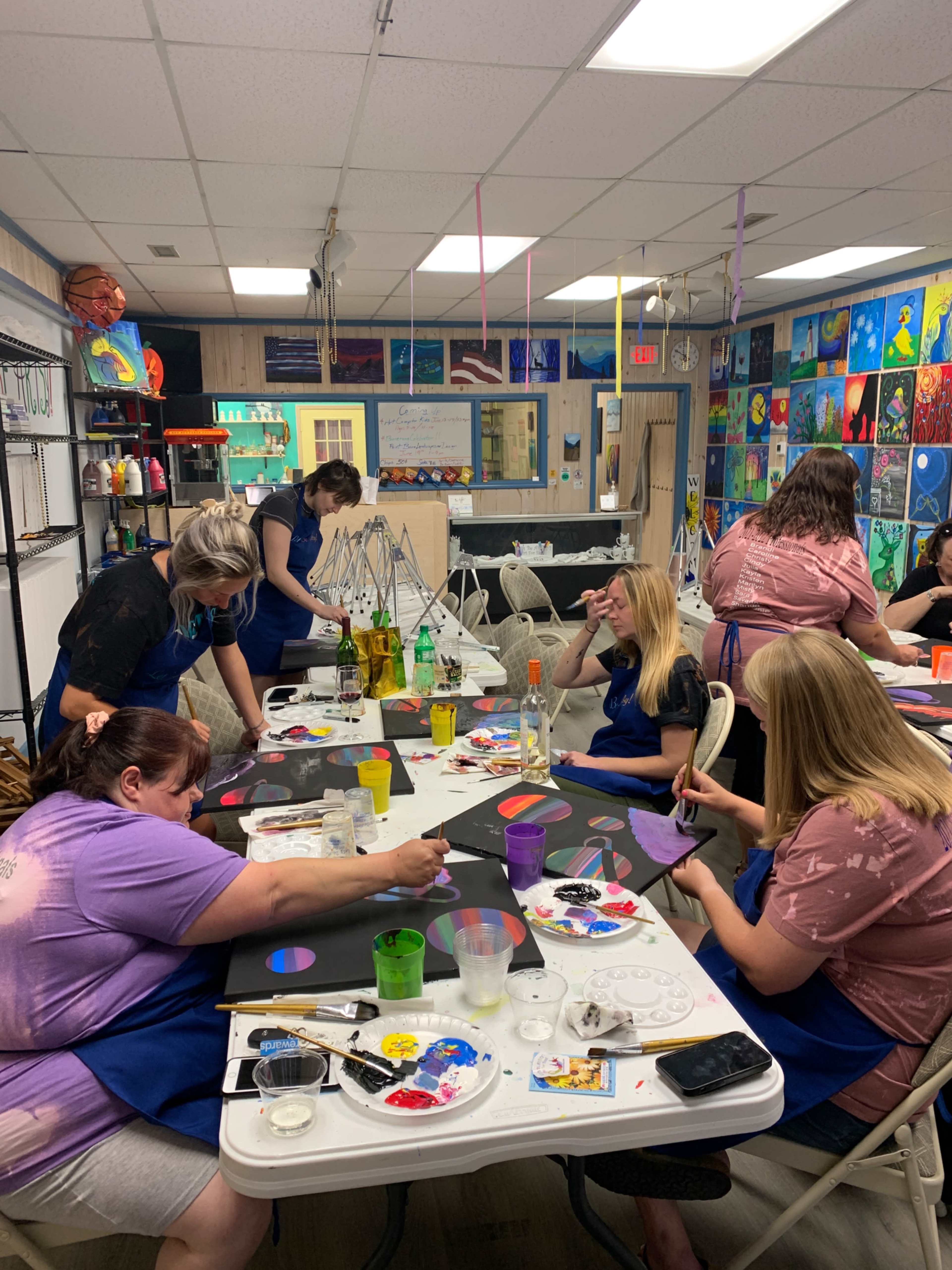 A group of individuals in blue aprons paint on canvases at a community art studio filled with colorful artwork on the walls.