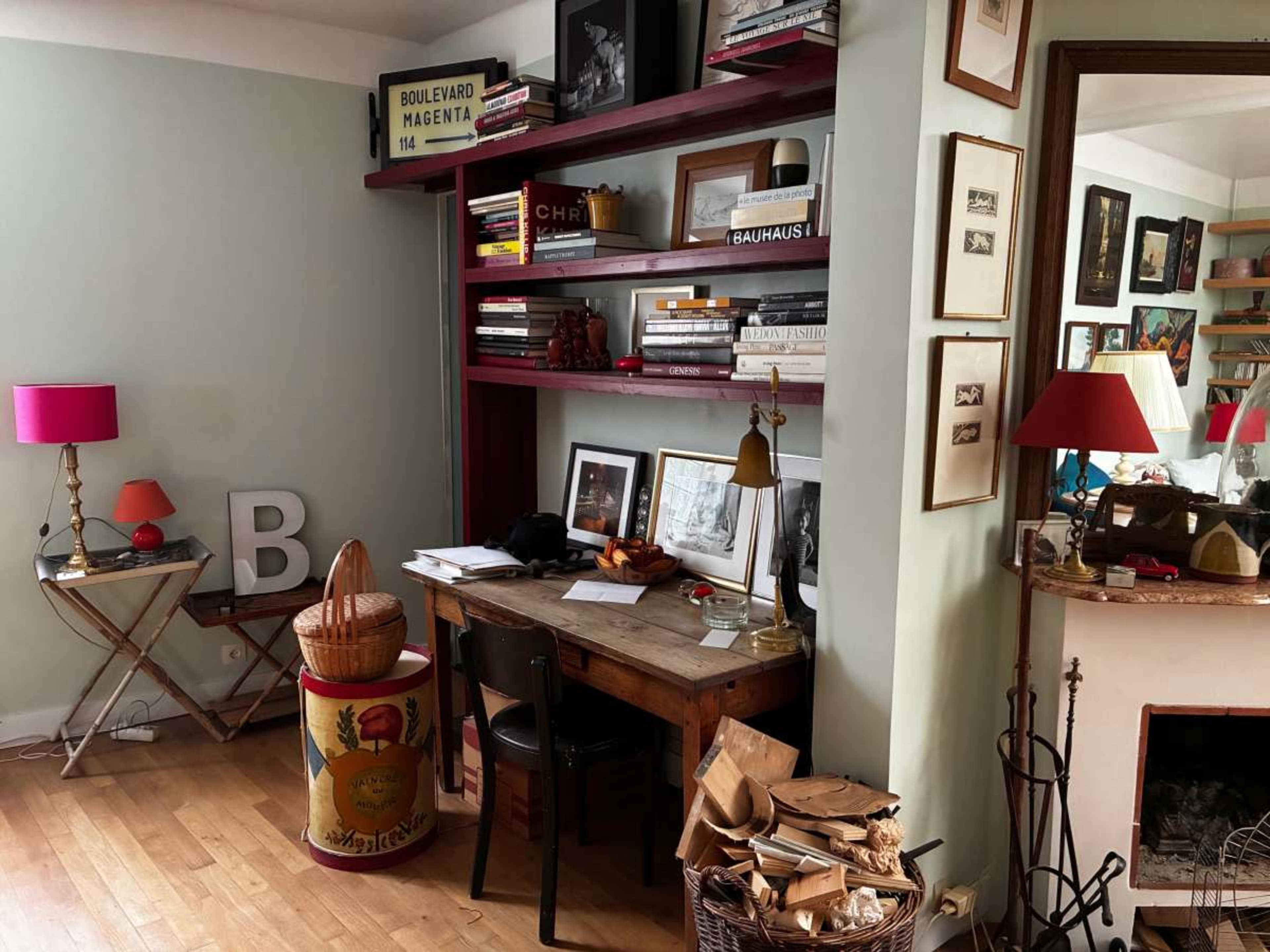 The image shows a cozy interior with a wooden desk, a bookshelf filled with books and framed photos, a decorative basket, and two red lamps in a room with hardwood flooring.