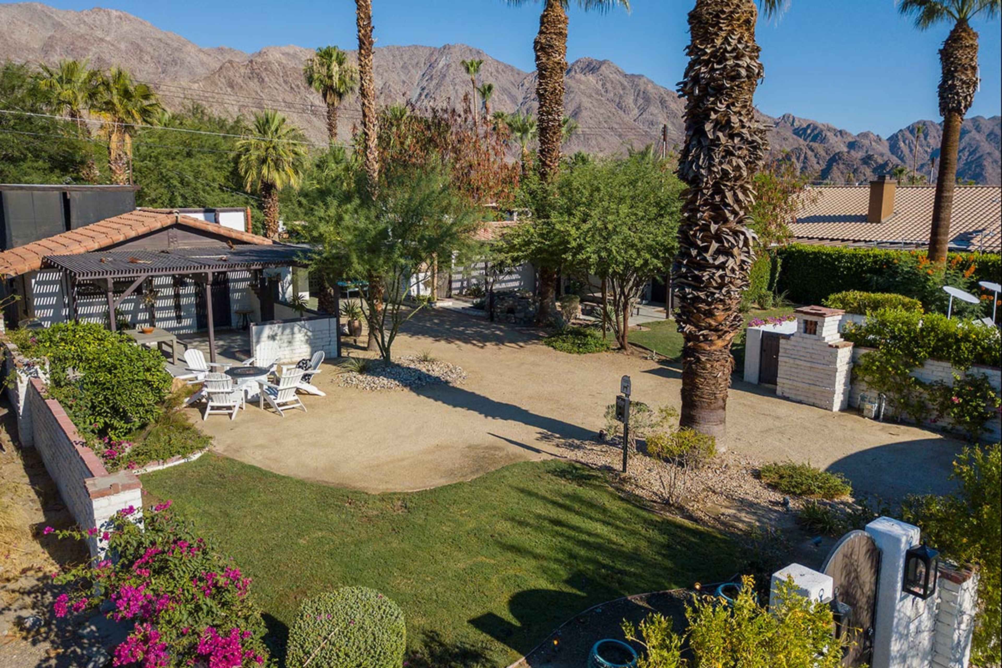 A landscaped outdoor area featuring palm trees, a gravel pathway, and a seating arrangement with white chairs near a small structure, all set against a backdrop of mountains.