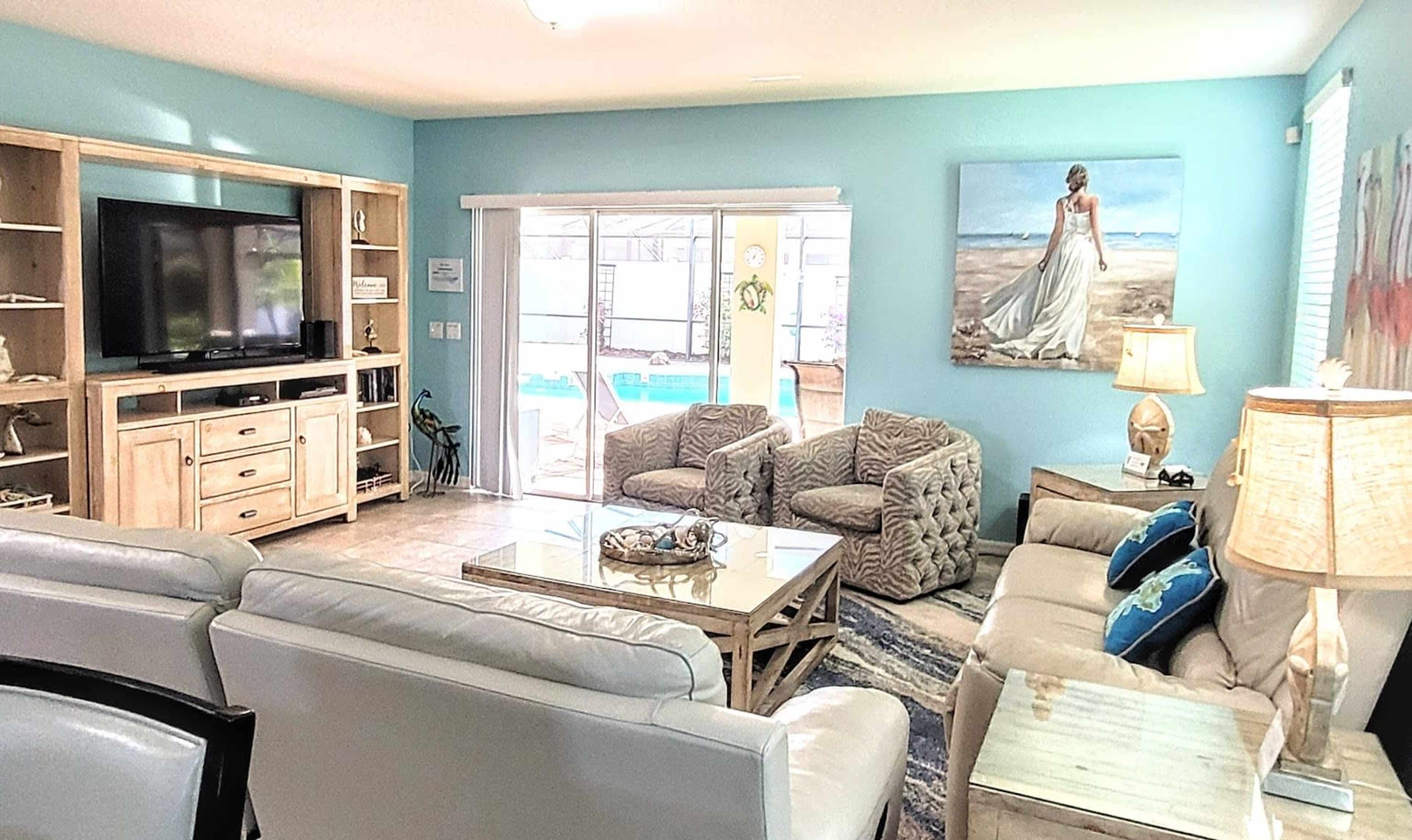 A modern living room with light-colored furniture, a large television, and a view of a pool area through sliding glass doors.
