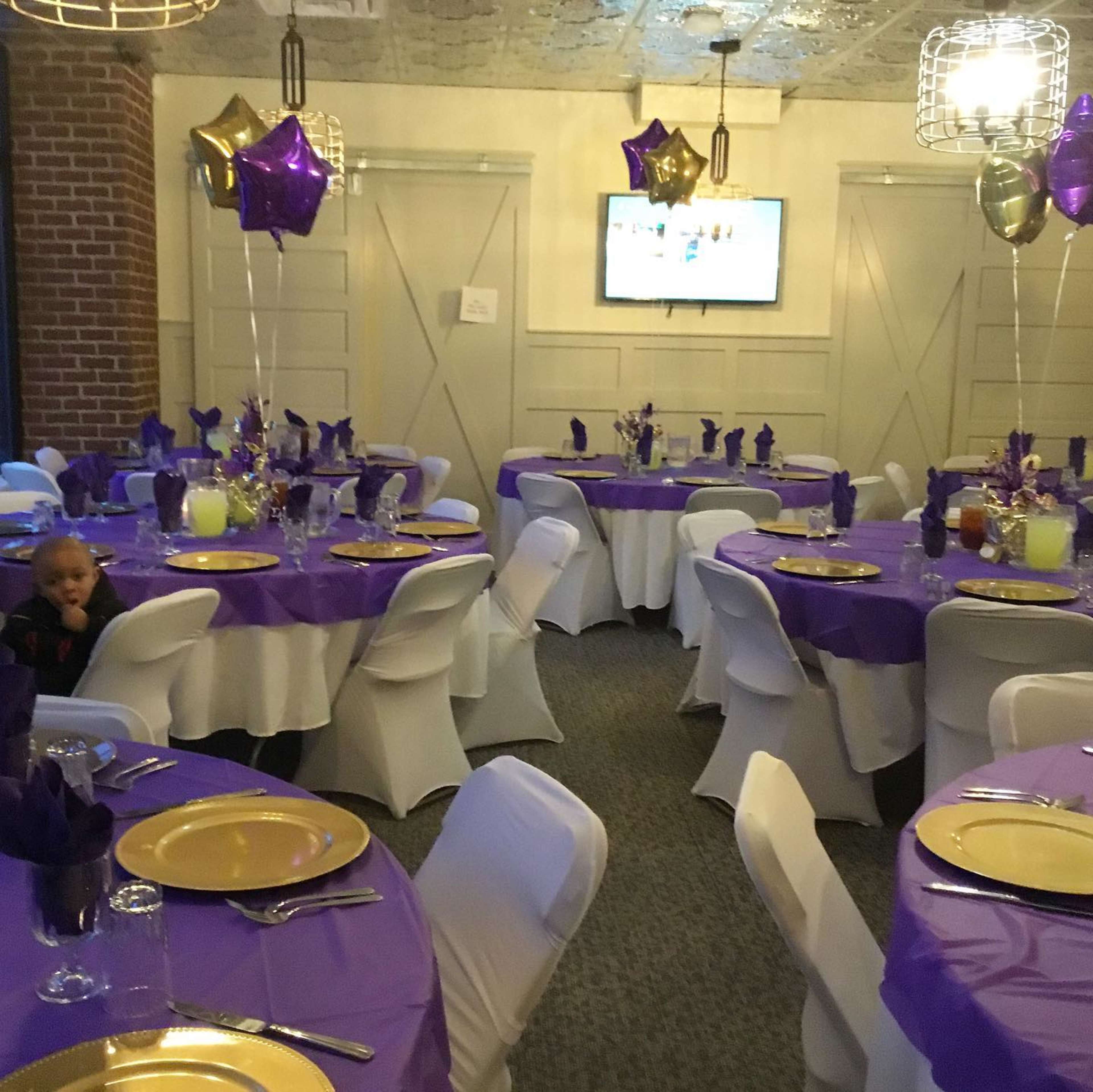 The image shows a banquet room with round tables set with purple tablecloths, gold plates, and decorative balloons, along with a television screen on the wall.