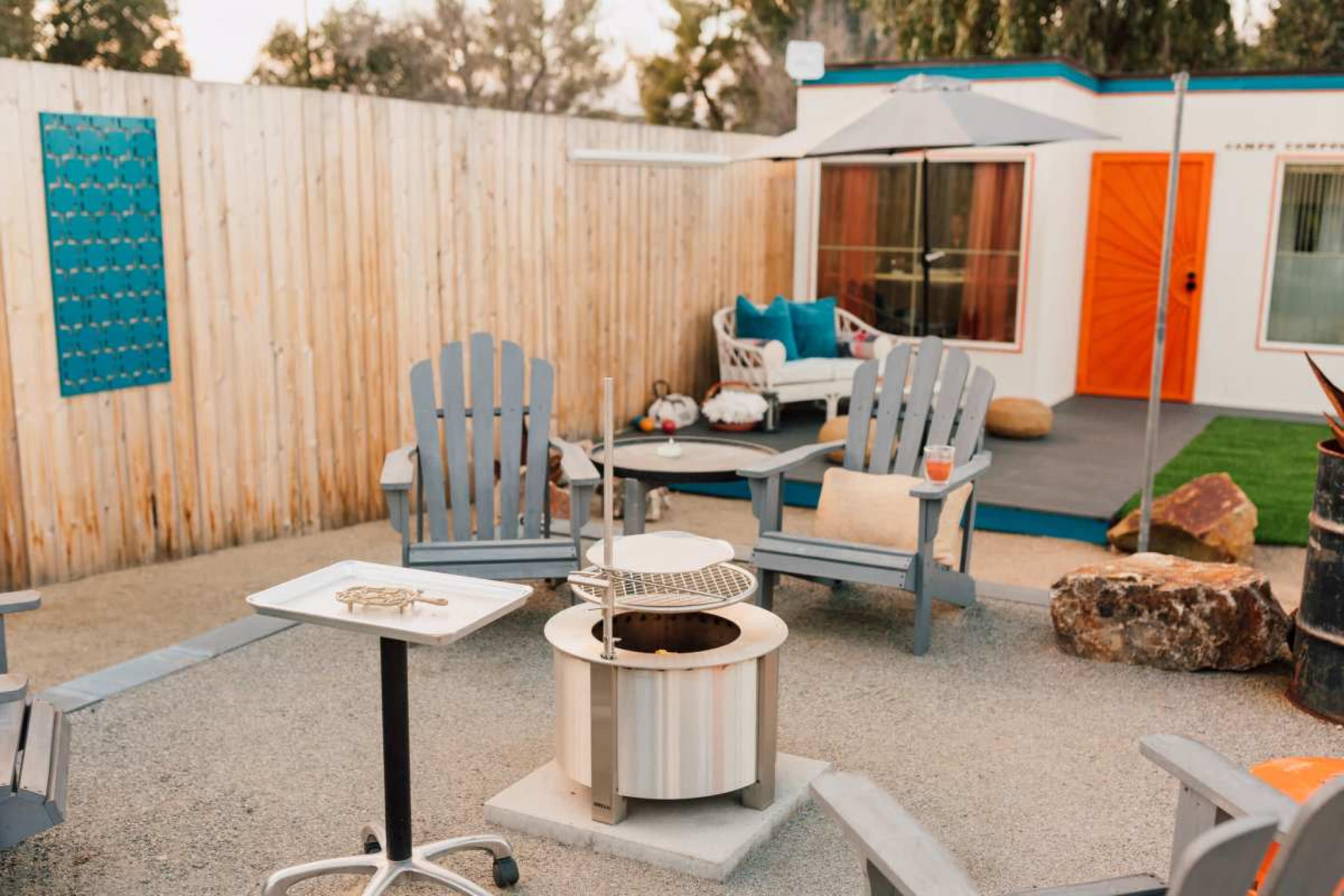 The image shows an outdoor seating area with several gray Adirondack chairs arranged around a circular fire pit, with a table and a small patio set in the background.