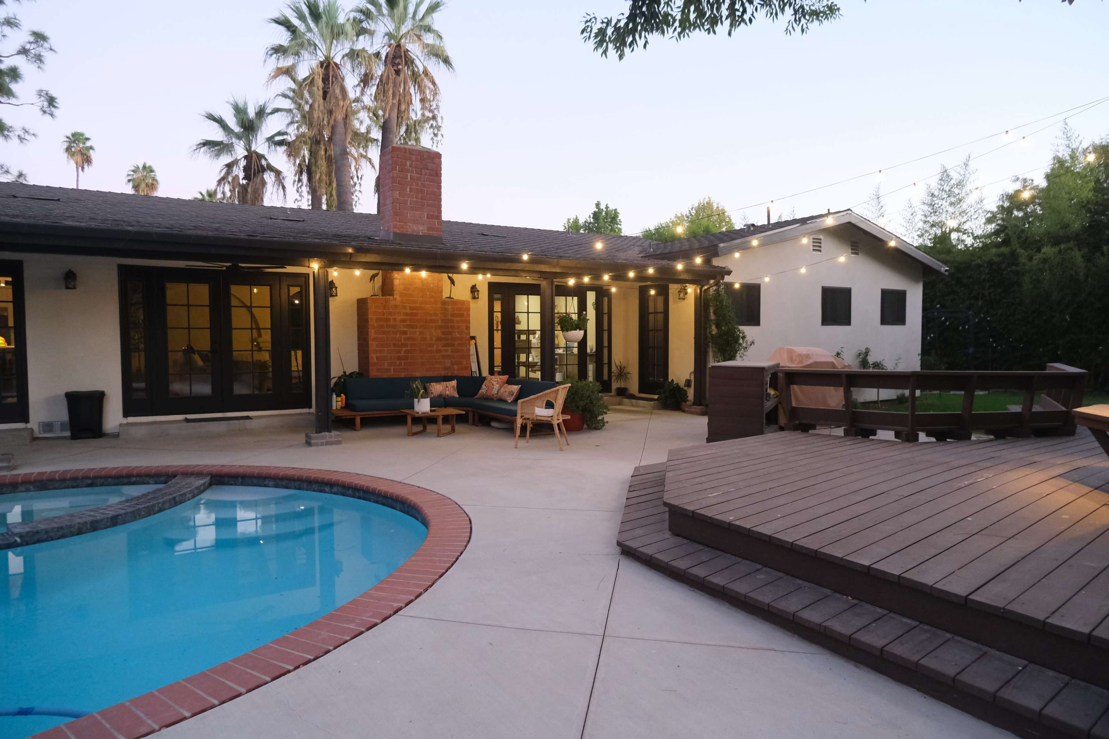 The image shows a backyard with a swimming pool, a wooden deck, and a single-story house surrounded by palm trees.