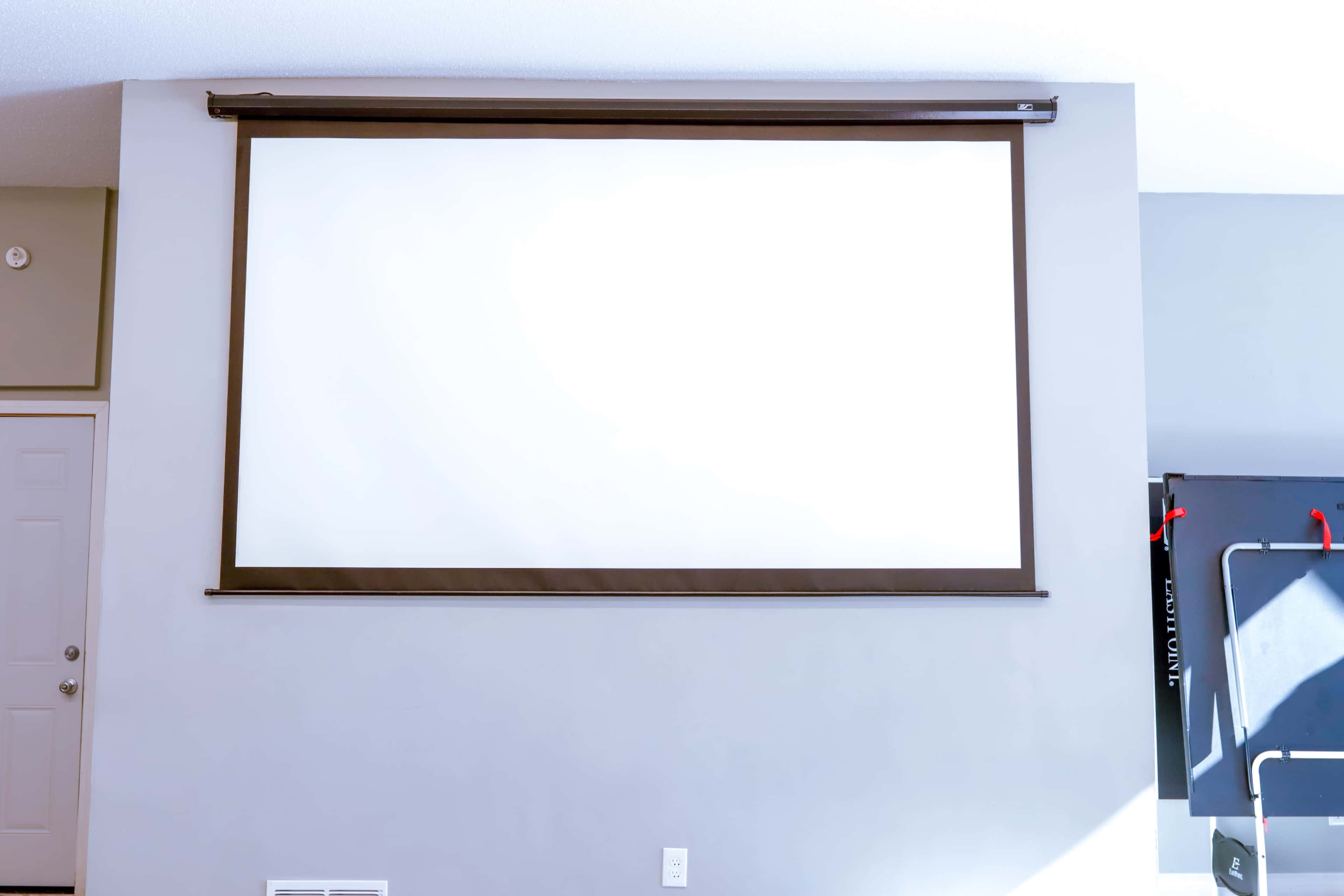 A large retractable projection screen mounted on a gray wall, with a door visible on the left and a piece of exercise equipment on the right.