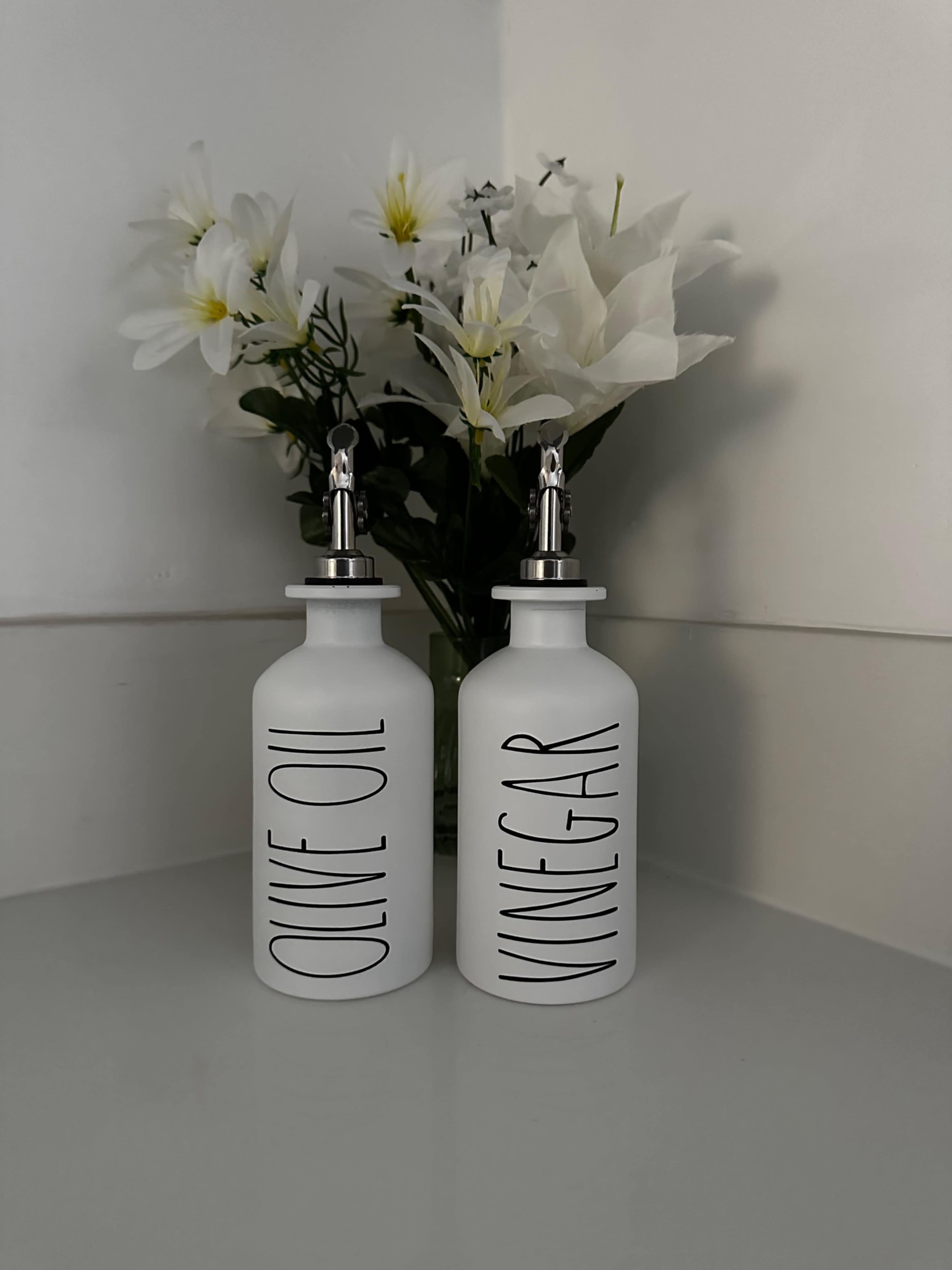 The image shows two labeled glass bottles for olive oil and vinegar beside a bouquet of white flowers on a white surface.