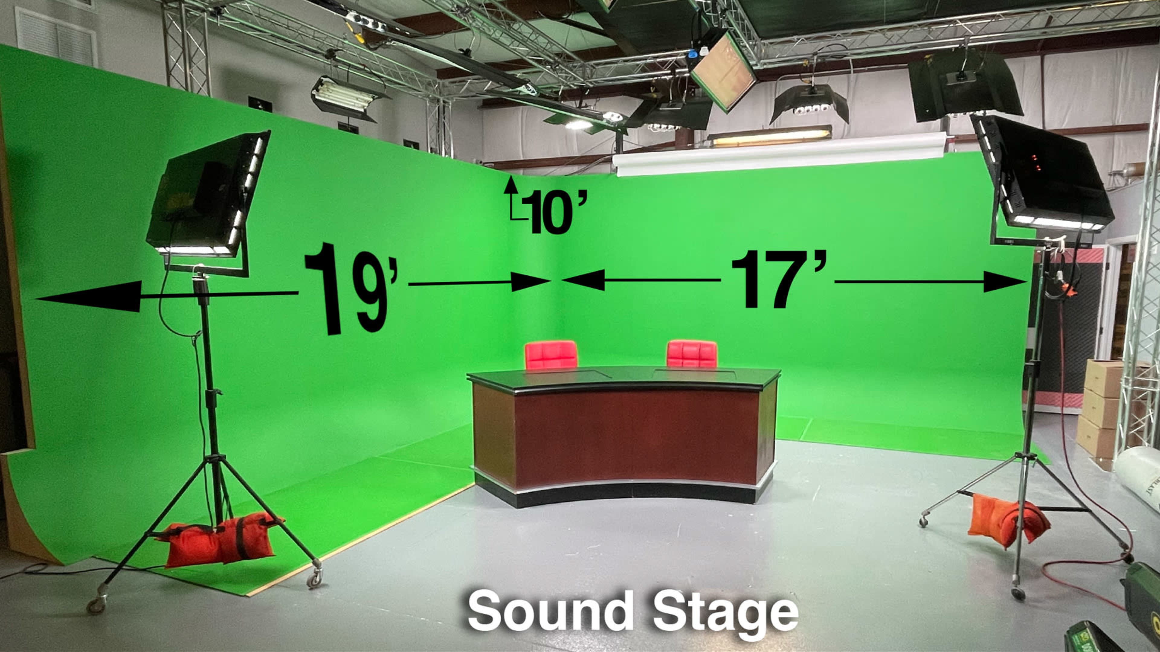 A sound stage with a green backdrop, a dark wooden desk, two red chairs, and lighting equipment set up.