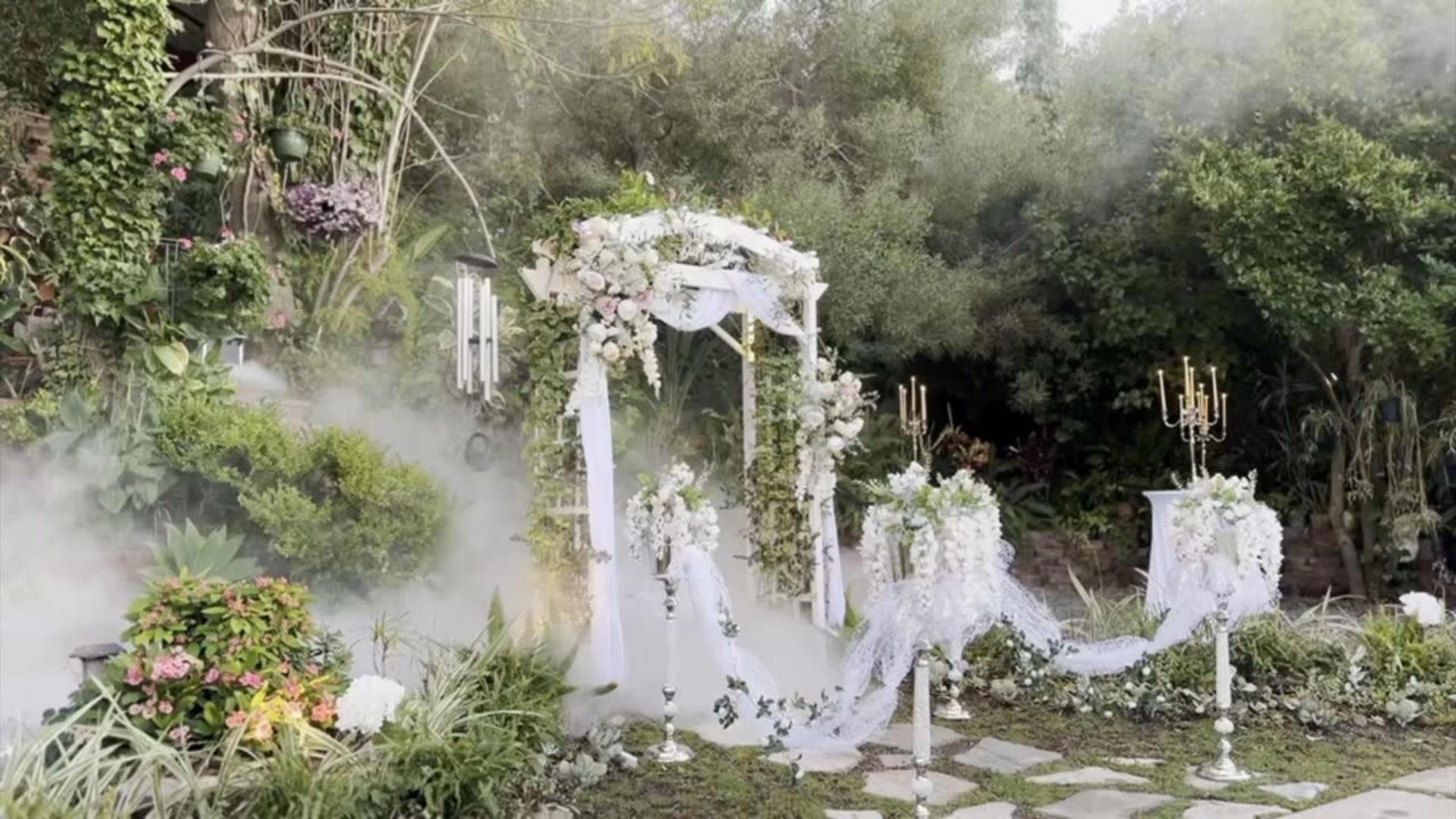 A decorative outdoor wedding altar adorned with white flowers and drapery is set against a lush green backdrop.