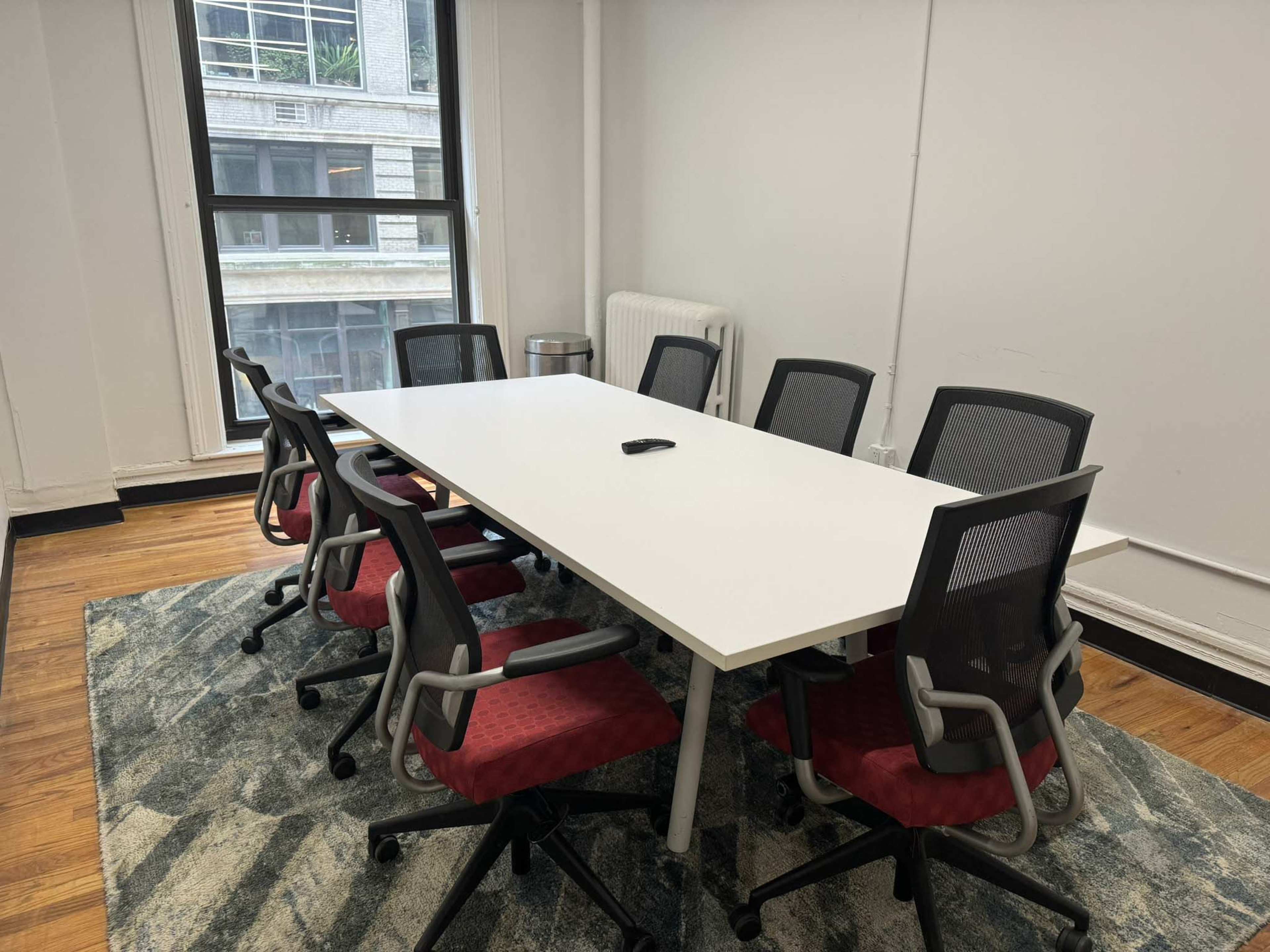 A bright conference room features a large white table surrounded by eight chairs on a patterned rug.
