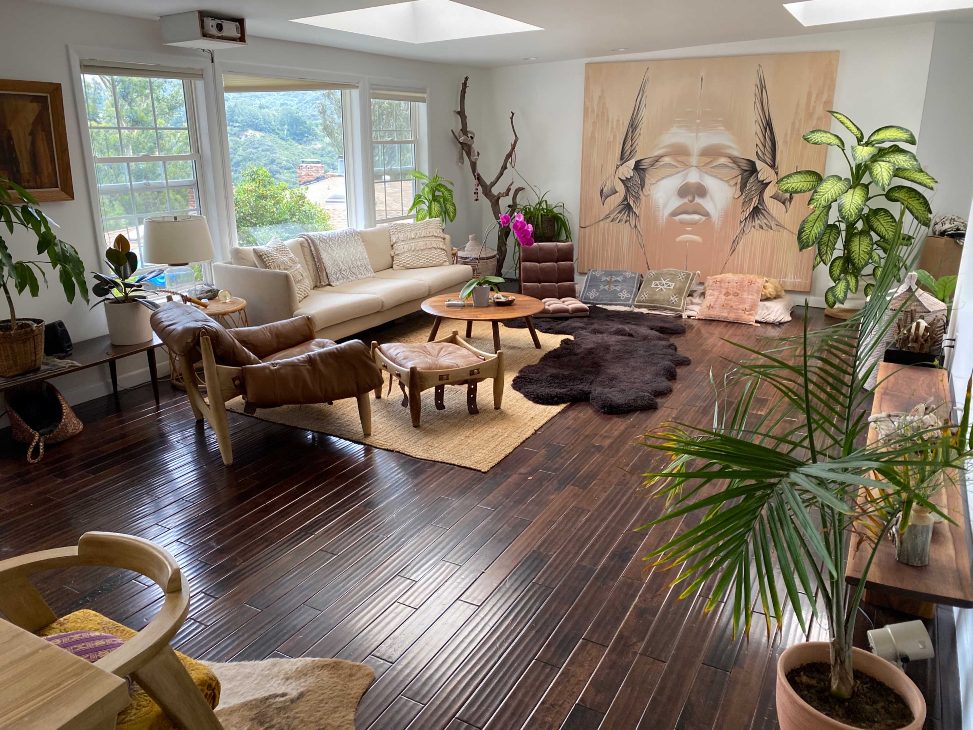 The room features a spacious sitting area with a large white sofa, leather chairs, a wooden coffee table, and an artwork depicting a face on the wall, surrounded by plants and natural light.