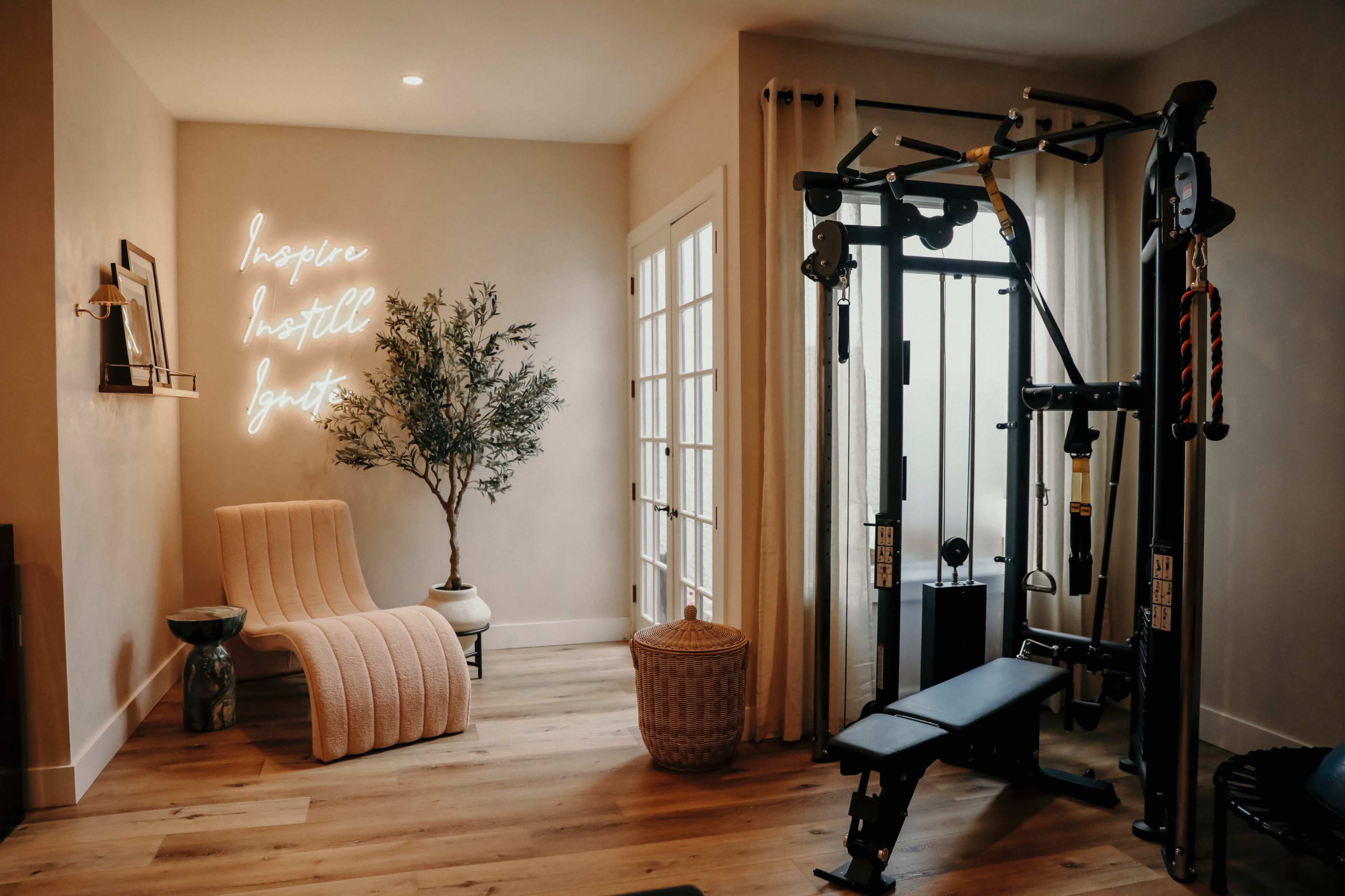 The image shows a modern home gym featuring workout equipment on one side and a cozy seating area with a neon sign on the wall.