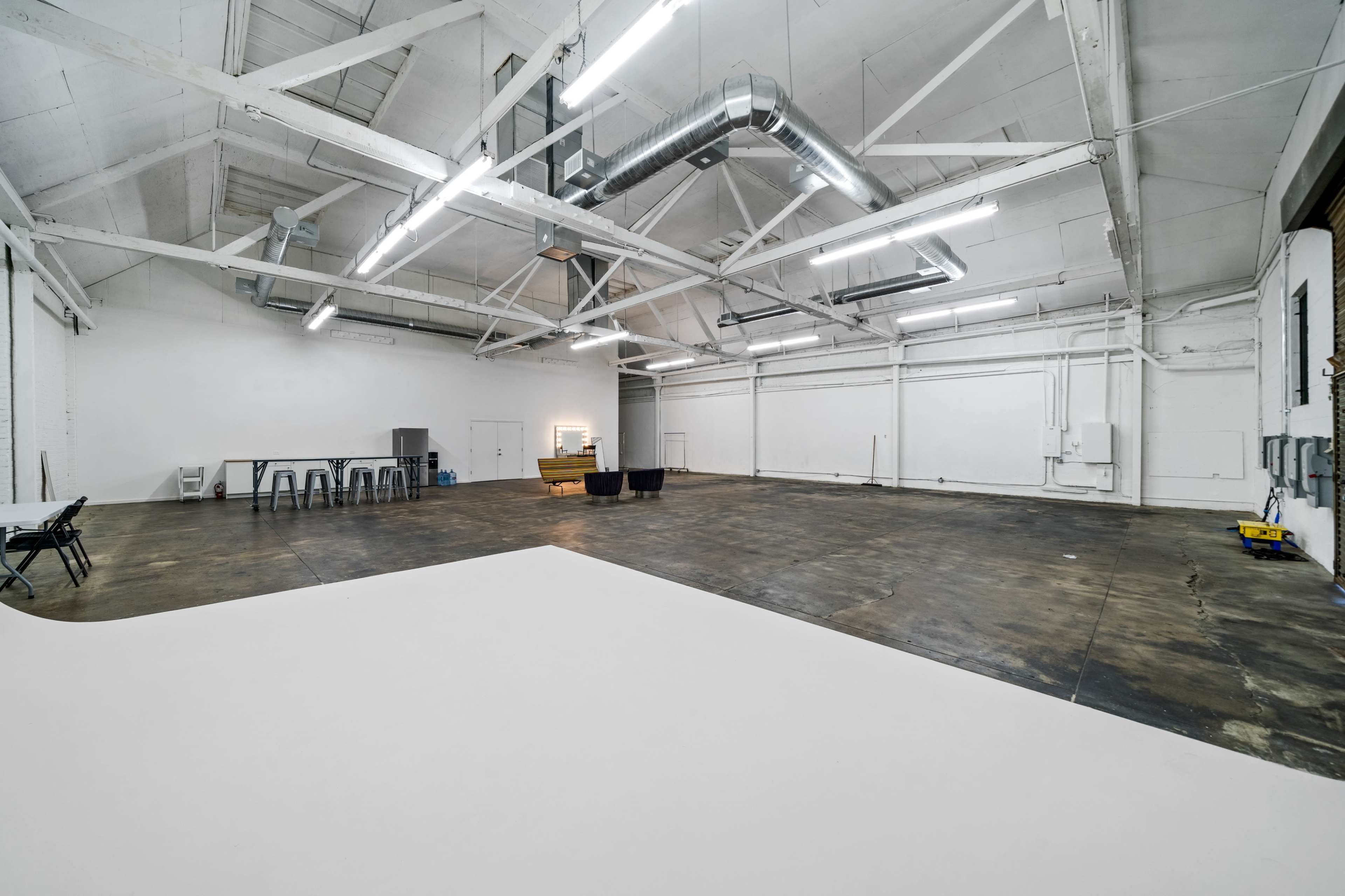 A spacious, empty industrial-style room with high ceilings, exposed beams, and large windows, featuring minimalist furnishings along one side.