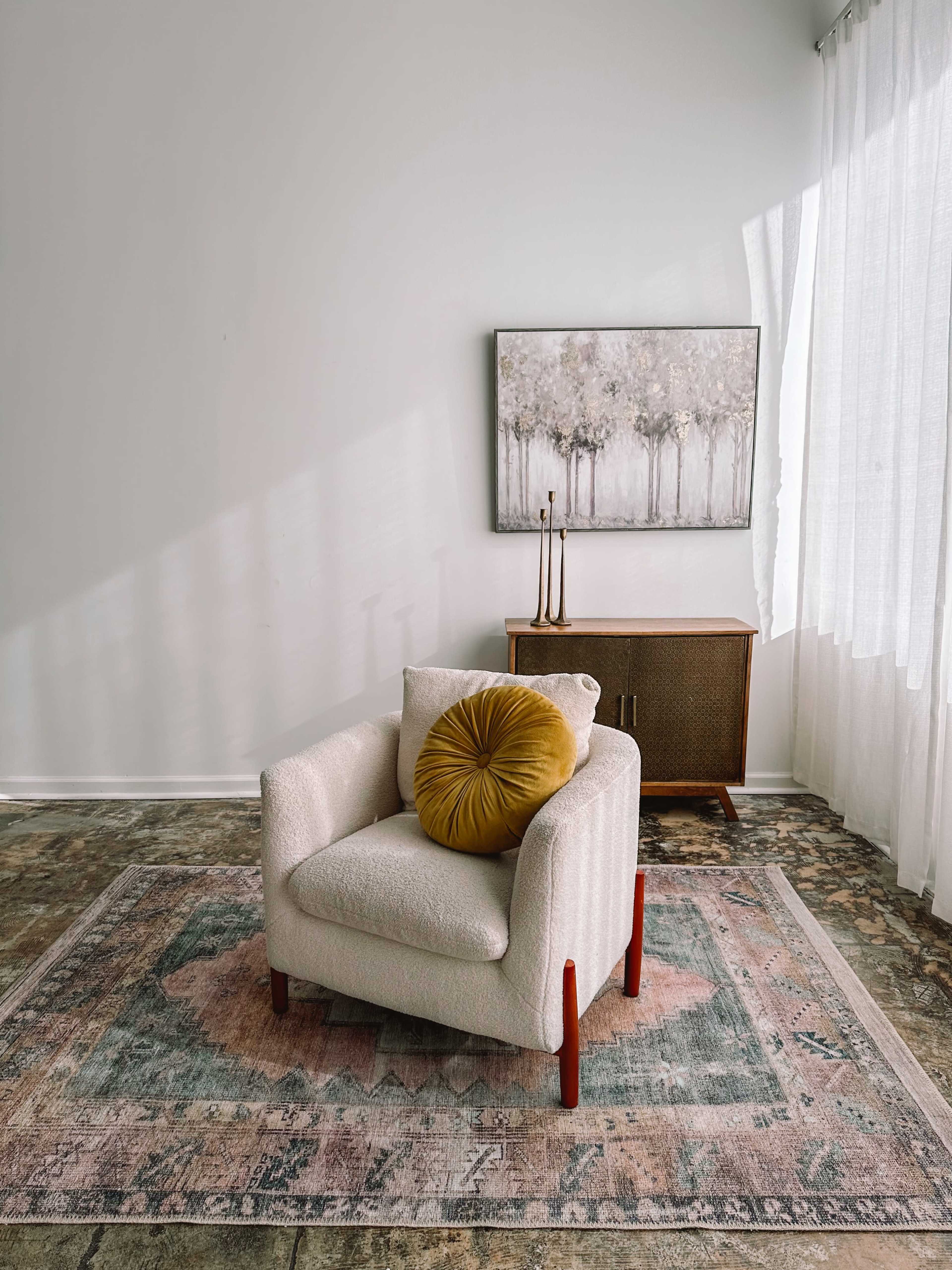 A single upholstered chair with a round cushion is positioned on a patterned rug in a room with large windows and a piece of artwork on the wall.