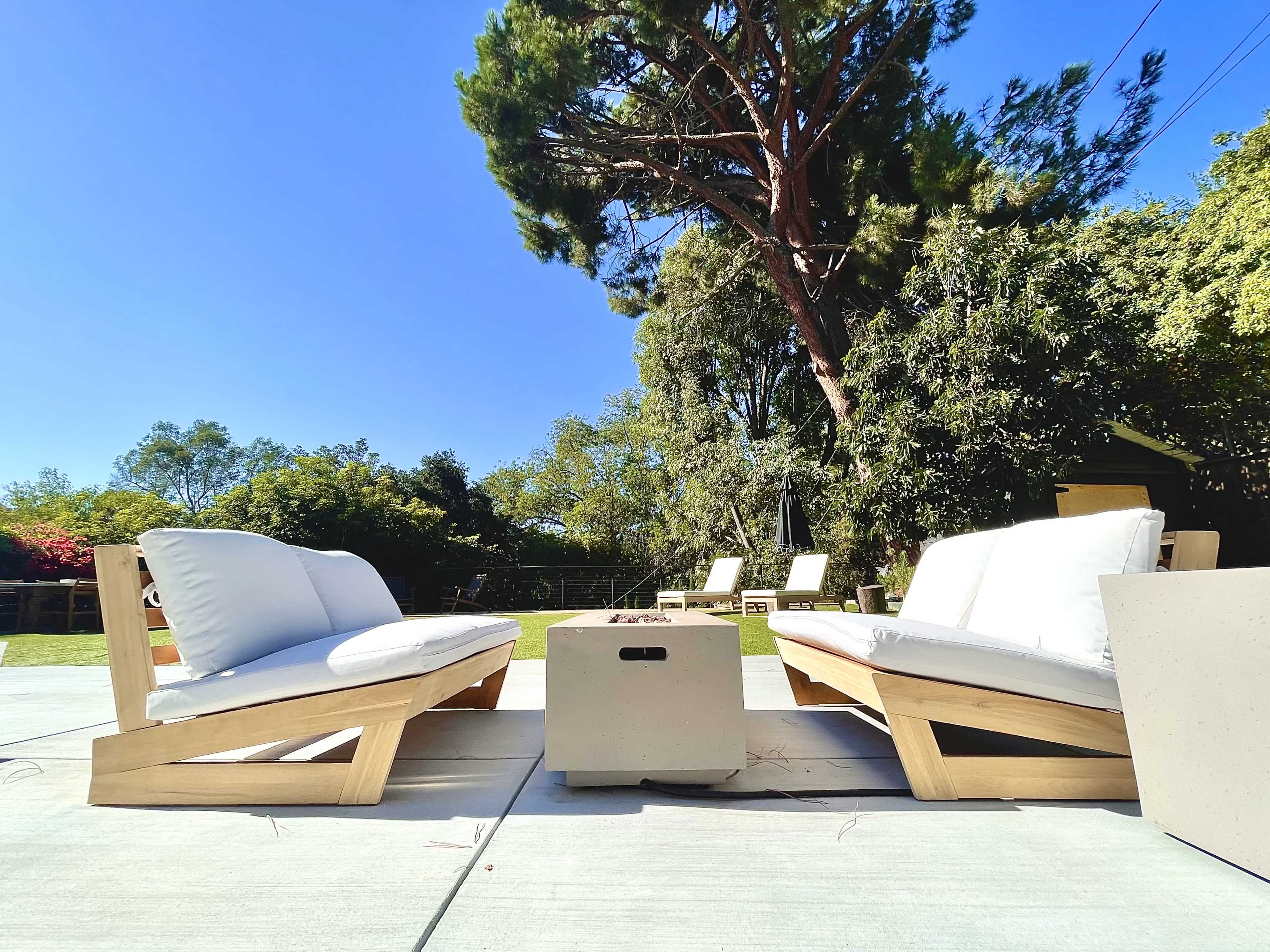 Two lounge chairs with white cushions are positioned facing each other around a concrete fire pit in a backyard setting with trees and blue sky in the background.