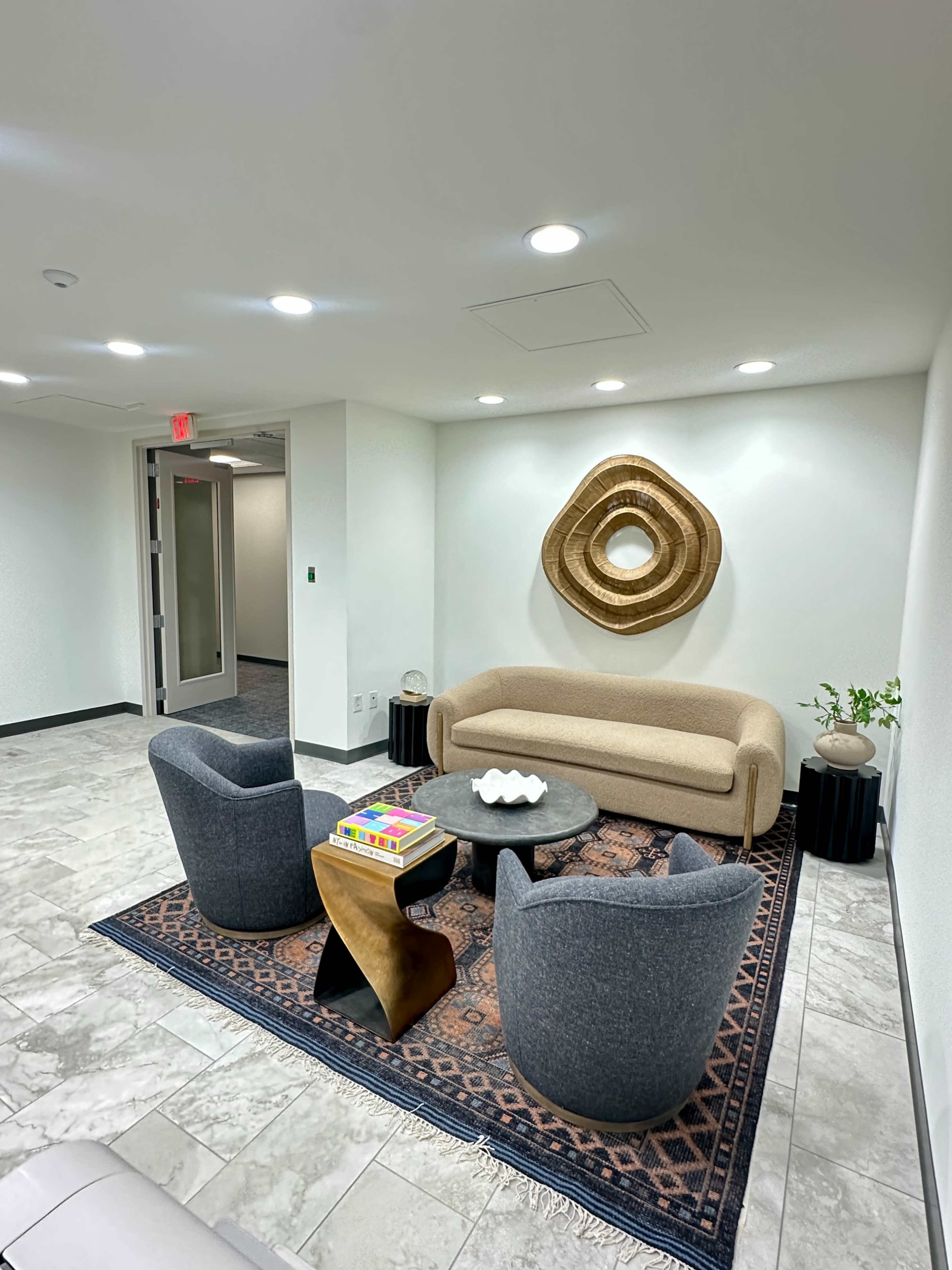 The image shows a modern office lounge area featuring a beige sofa, two gray armchairs, a round black table, and a decorative wall piece.