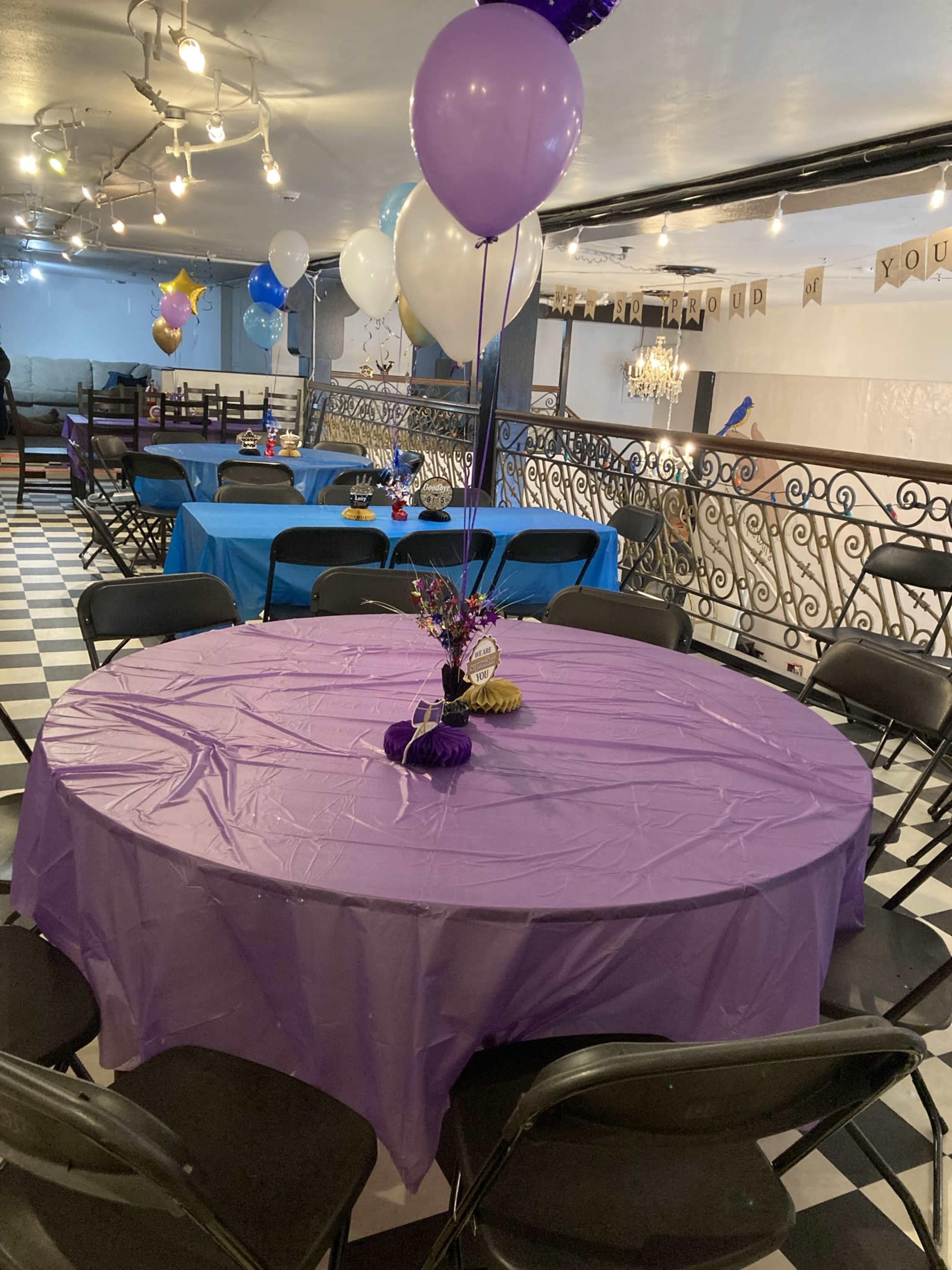 The image shows a party venue with round purple tables, each adorned with a centerpiece, surrounded by black chairs under string lights.