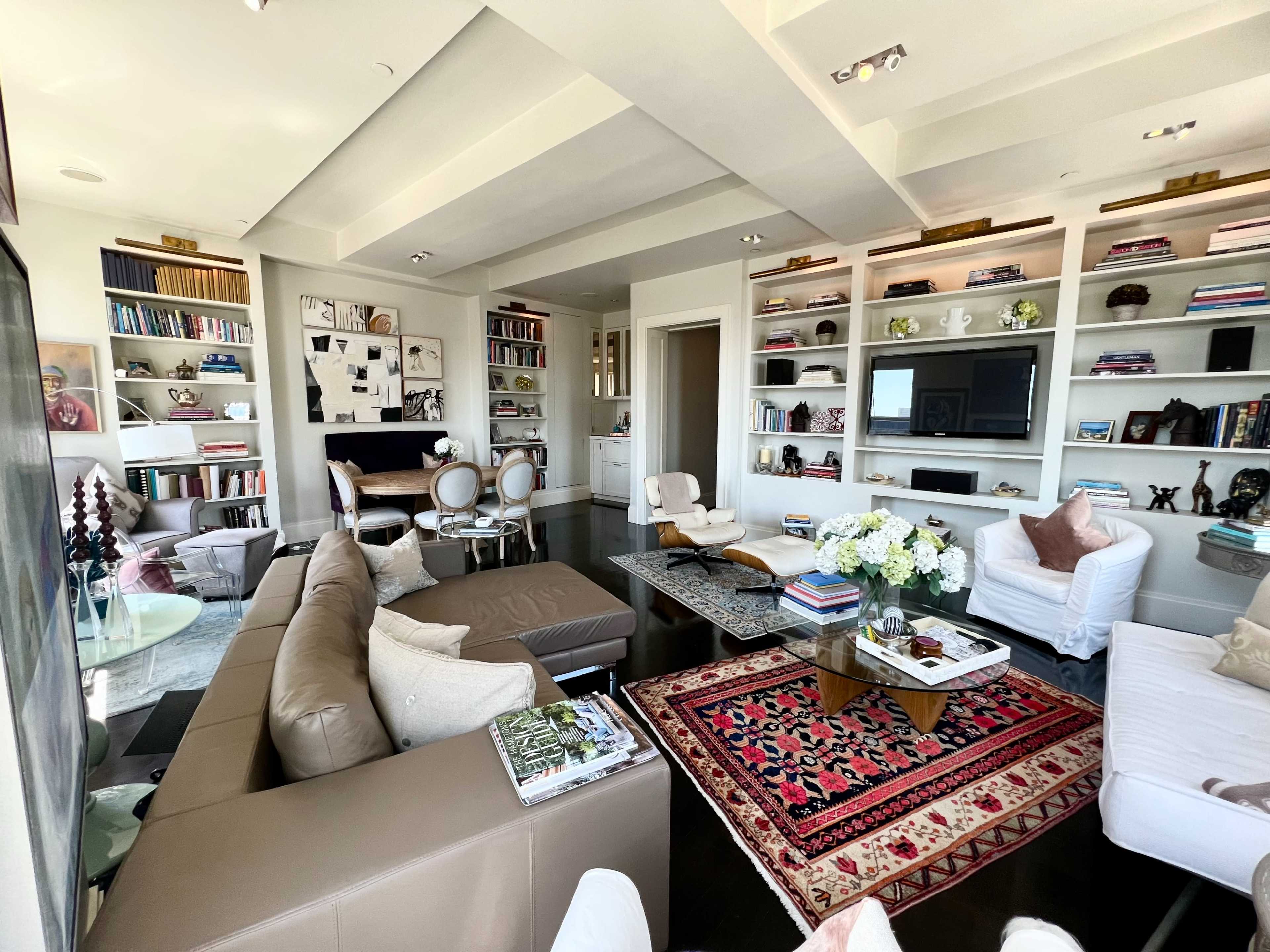 A modern living room with a sectional couch, a white chair, bookshelves filled with books and decor, and a coffee table adorned with flowers and magazines.