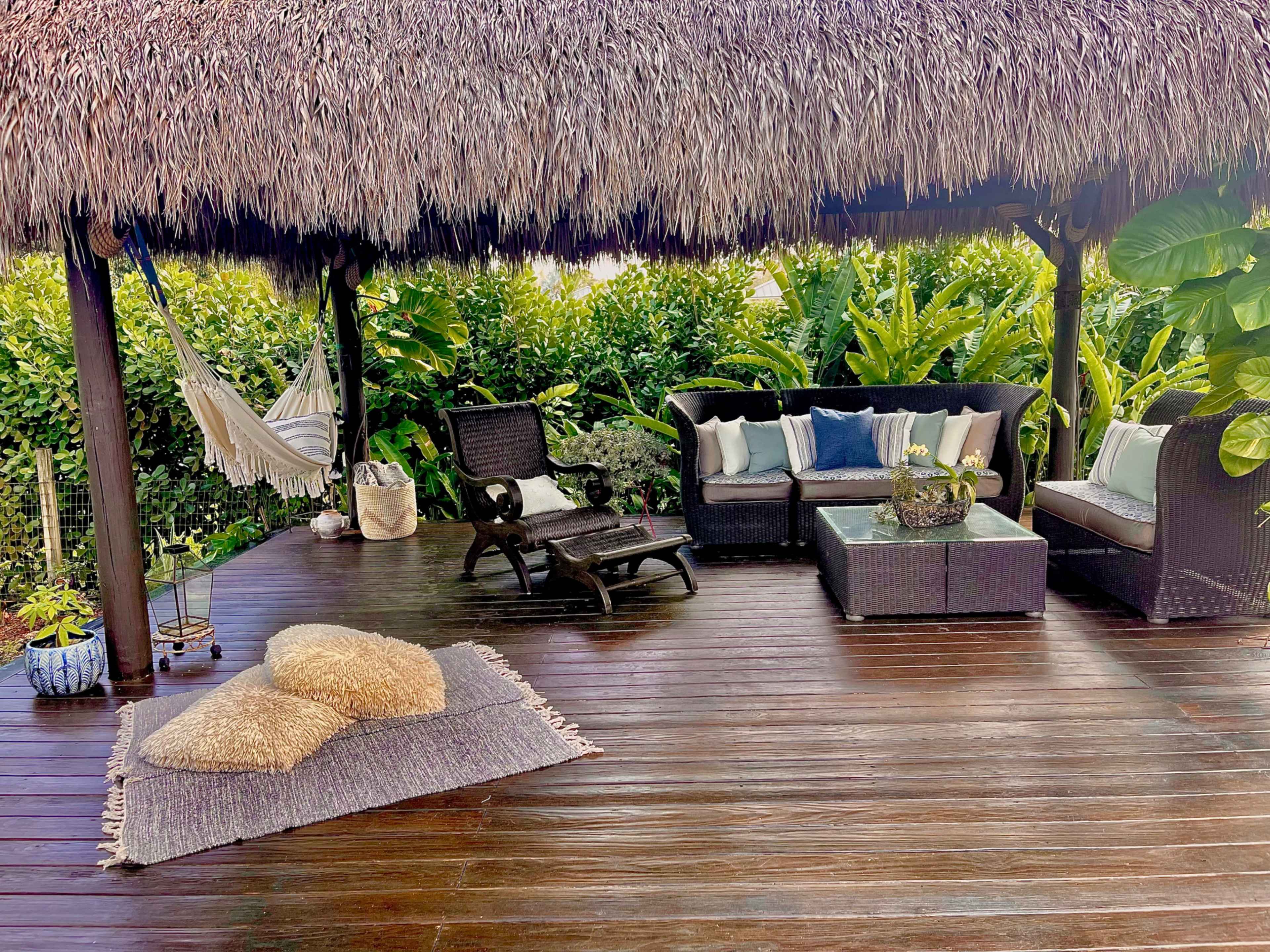 A shaded patio area with a thatched roof, featuring a seating arrangement, a hammock, and decorative plants.